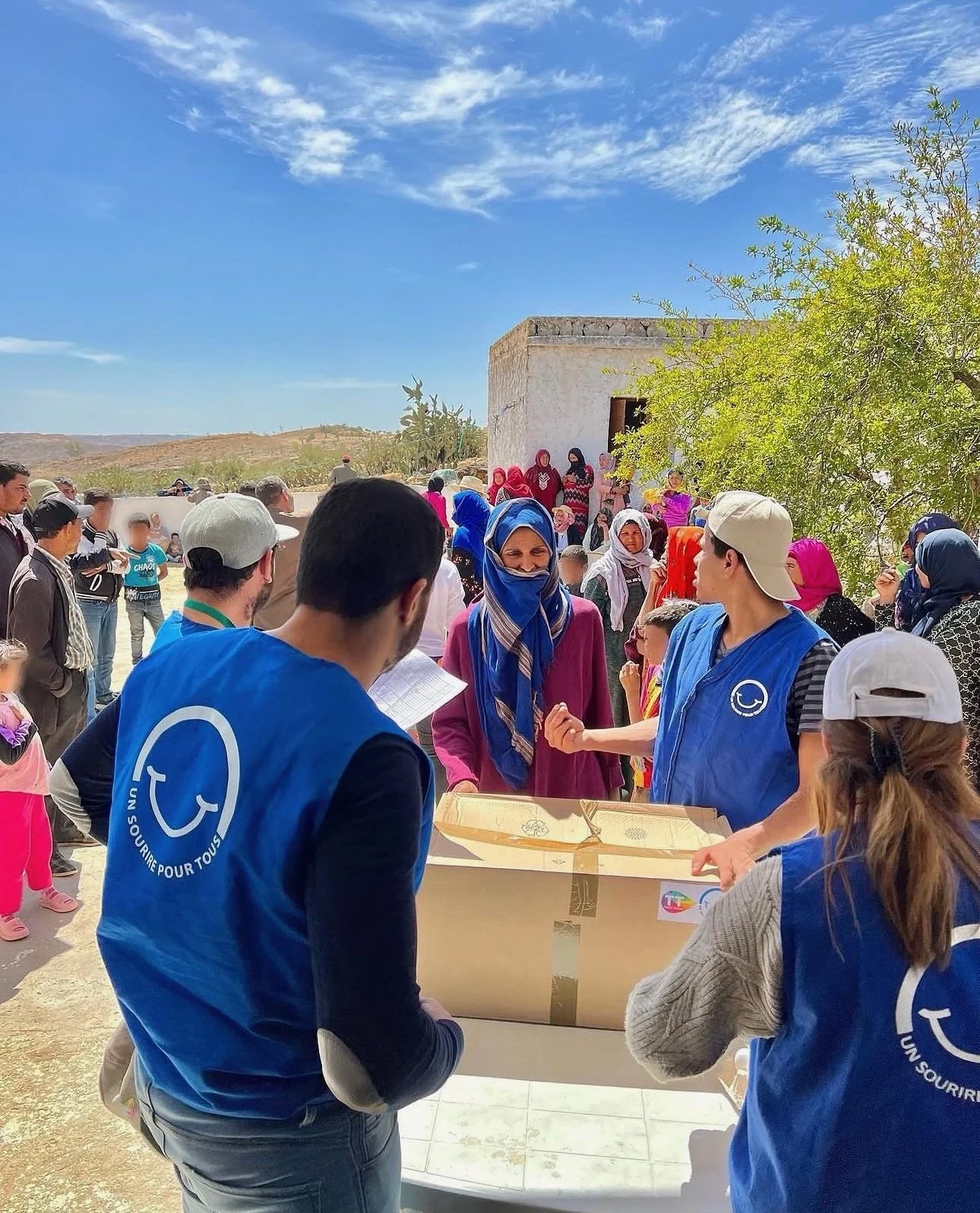 A group of volunteers and local villagers gather outdoors under a bright blue sky with clouds. Volunteers wearing blue vests with a logo and text are engaging with women and children, possibly distributing aid or supplies, near a small building and a green tree in a rural setting.