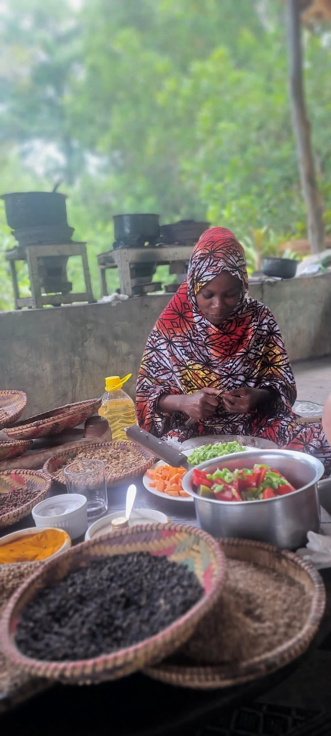 A woman wearing a colorful headscarf preparing vegetables at an outdoor food stall. Various bowls of vegetables, spices, and cooking ingredients are on the table, with a background of green foliage and cooking pots.