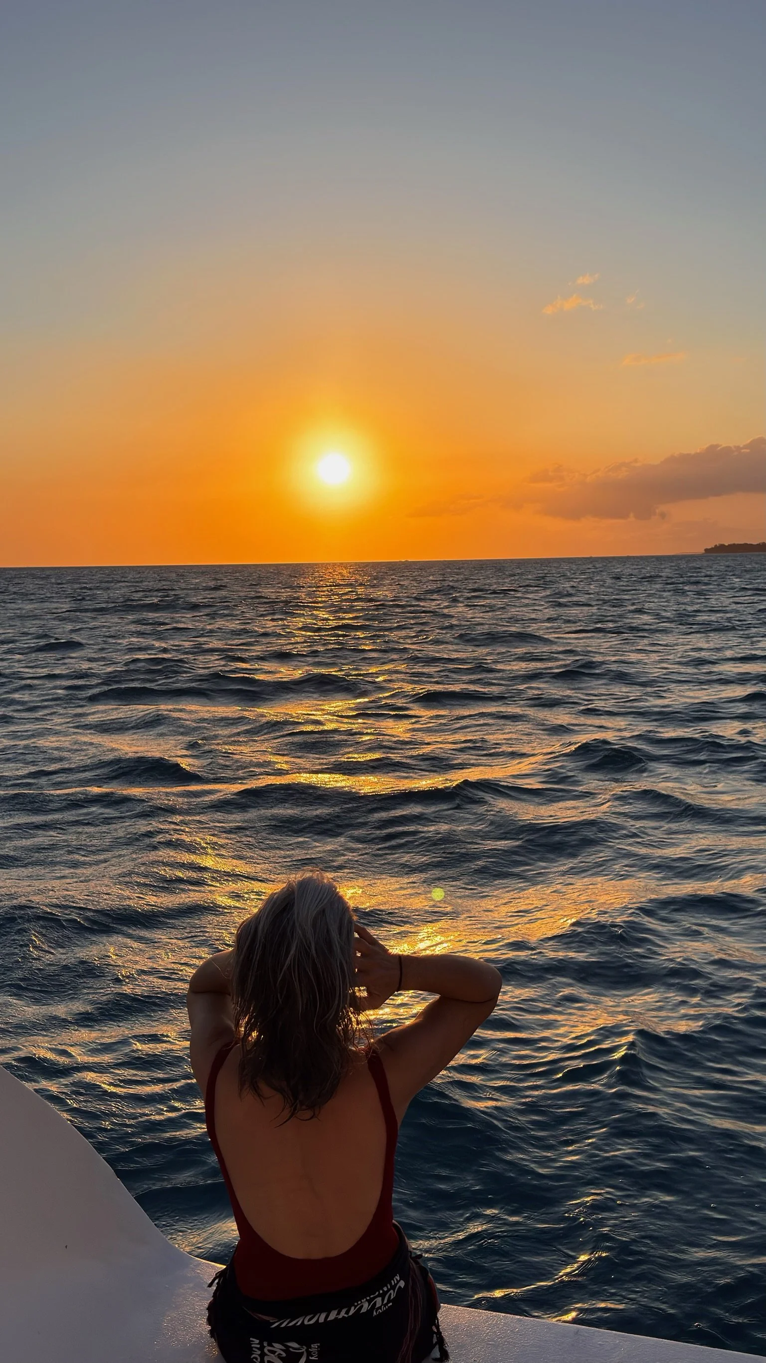 A woman with long hair wearing a red swimsuit, sitting on the edge of a boat, looking at an ocean sunset with orange, yellow, and pink hues in the sky and waves reflecting the sunlight.