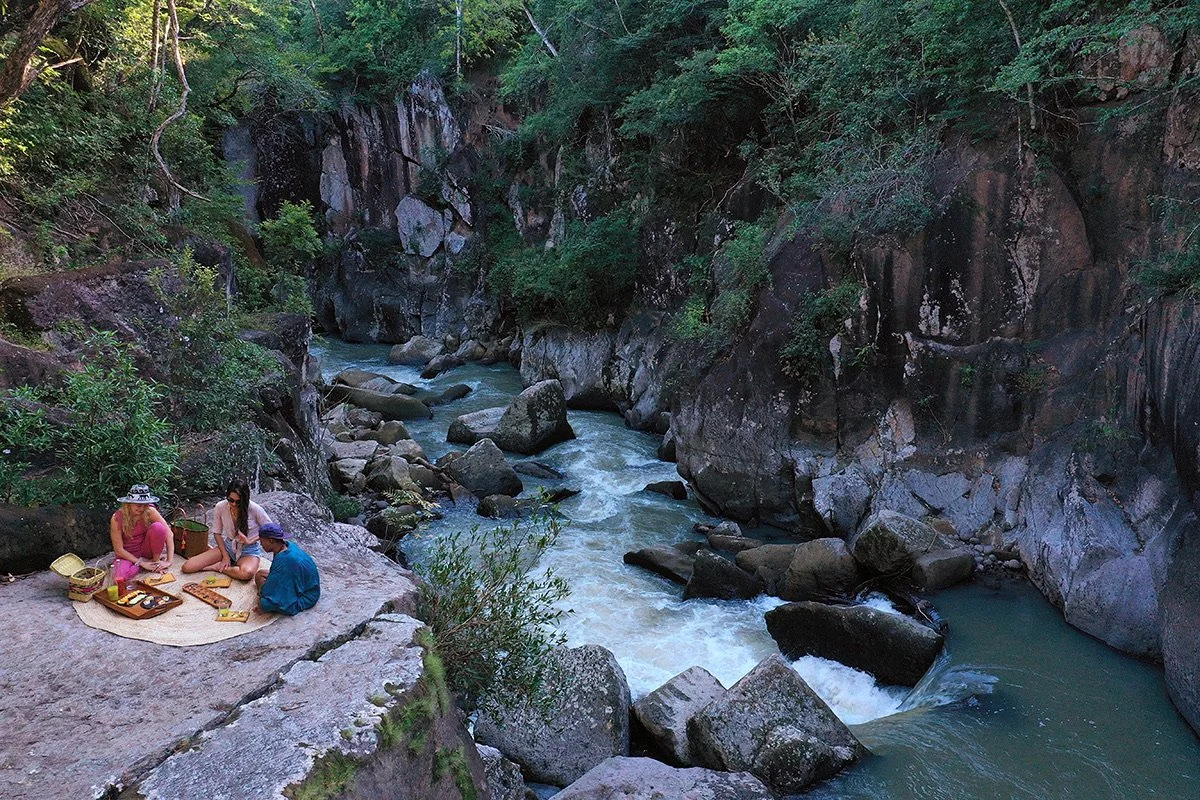 Three women are having a picnic on a large rock by a river in a forested canyon with tall cliffs and boulders.