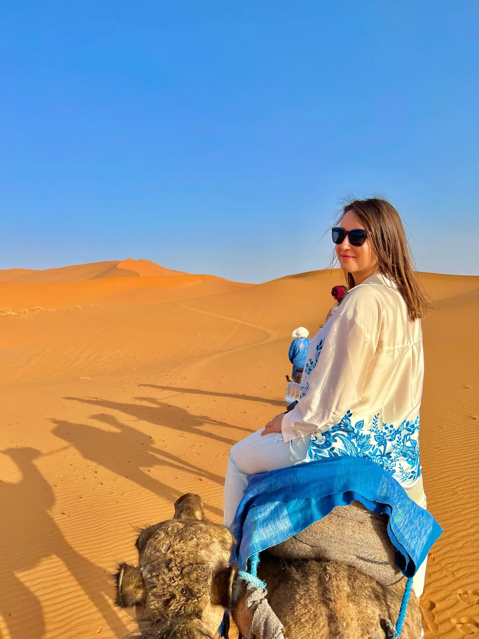 A woman wearing sunglasses and a white and blue embroidered dress sitting on a rock in a desert with sand dunes and a clear blue sky in the background.