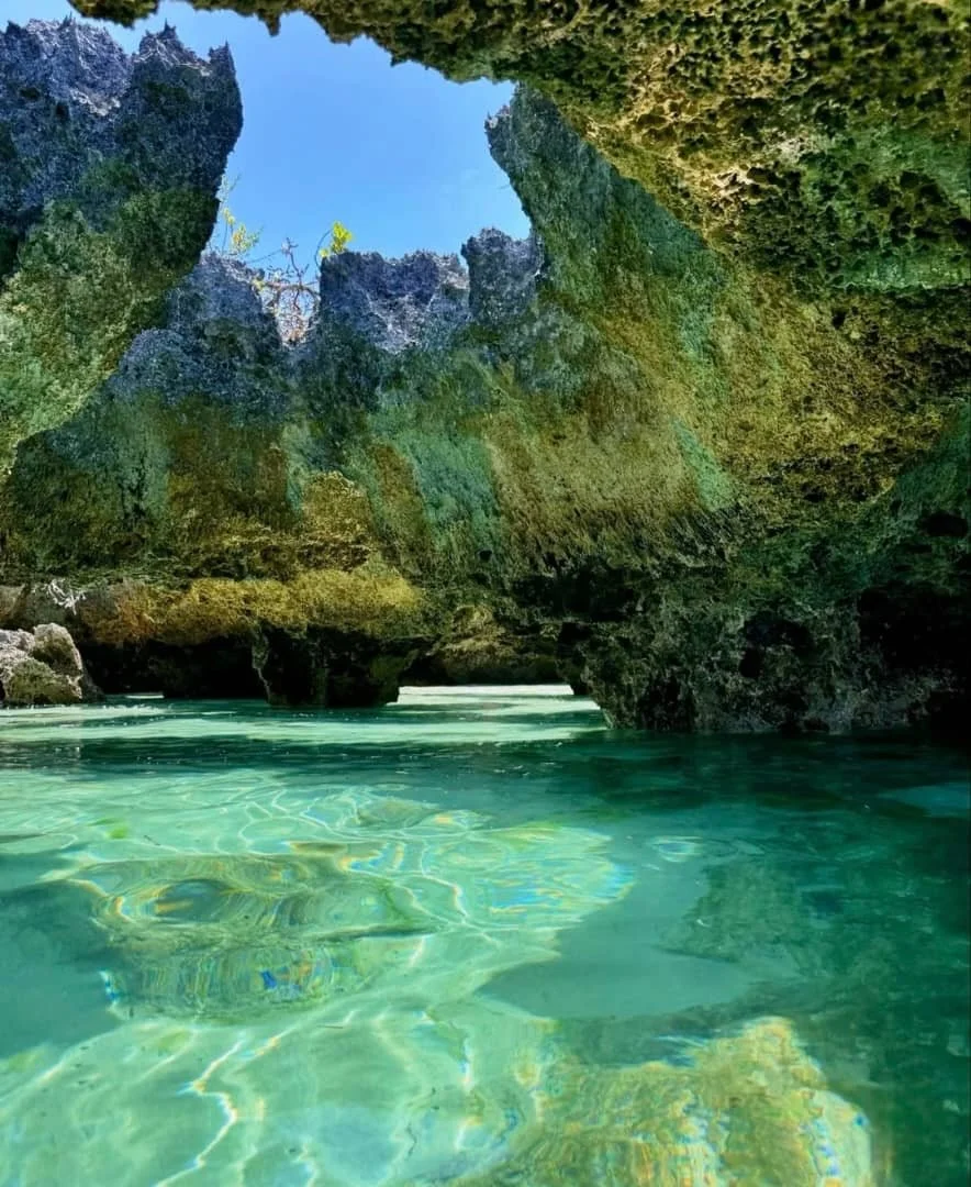Clear turquoise water at the base of a rocky coastal cliff, with the sky visible through the opening at the top.
