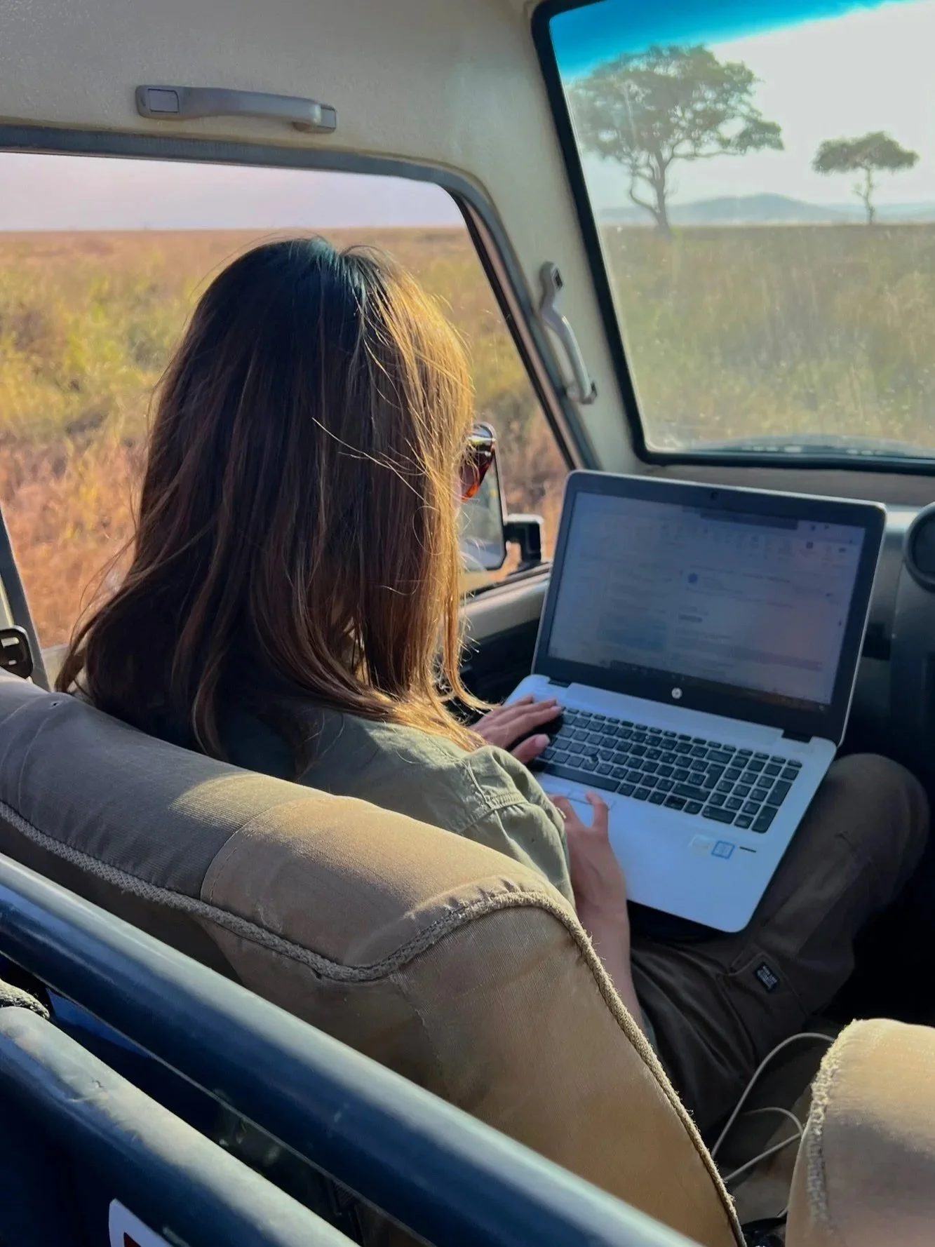 Woman working on a laptop in a vehicle traveling through a rural landscape with trees and open fields.