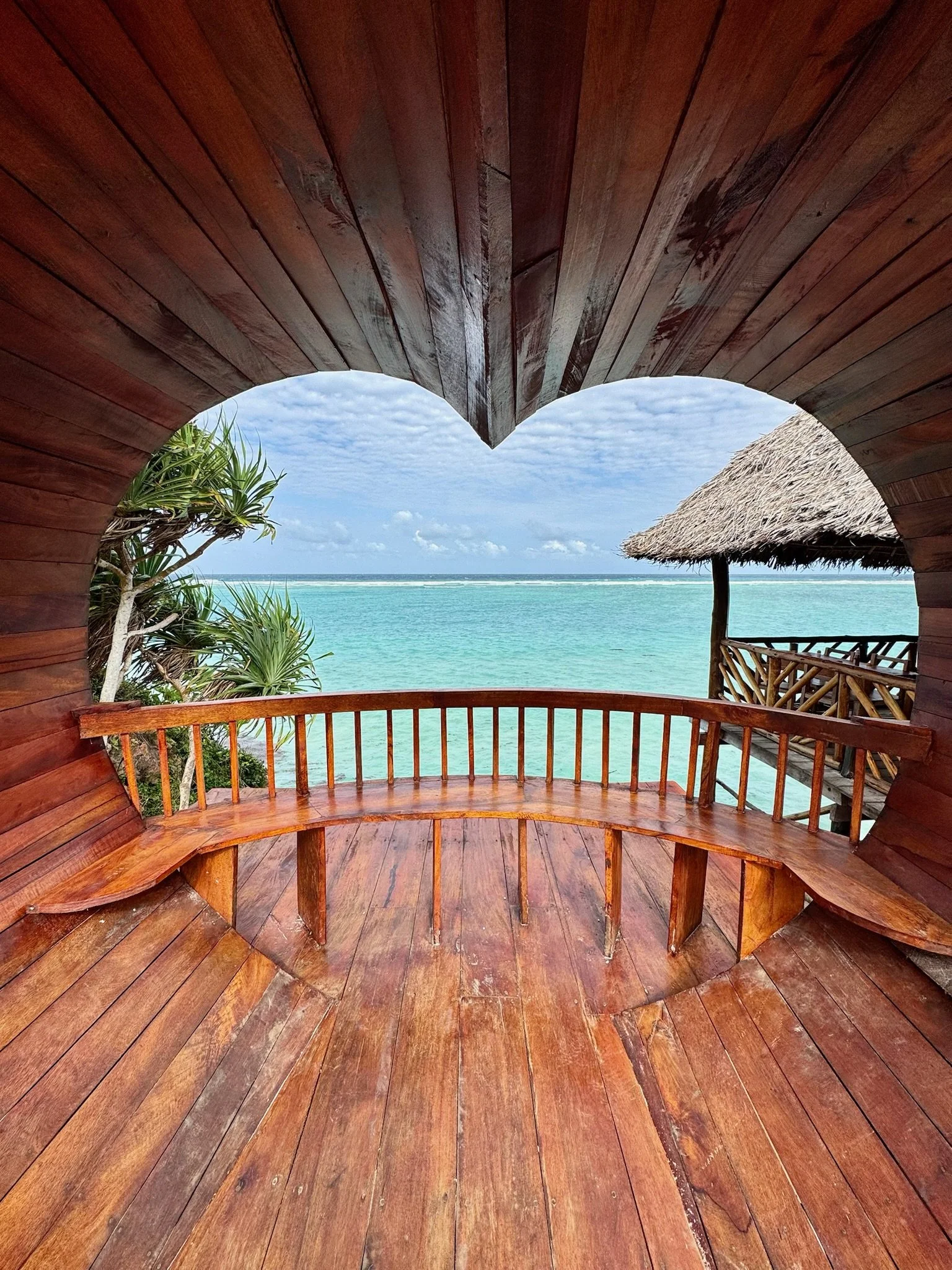 A view of a tropical beach scene, seen through a wooden archway with a curved bench, overlooking the clear turquoise sea, with a thatched-roof hut and some tropical plants on a sunny day with partly cloudy sky.