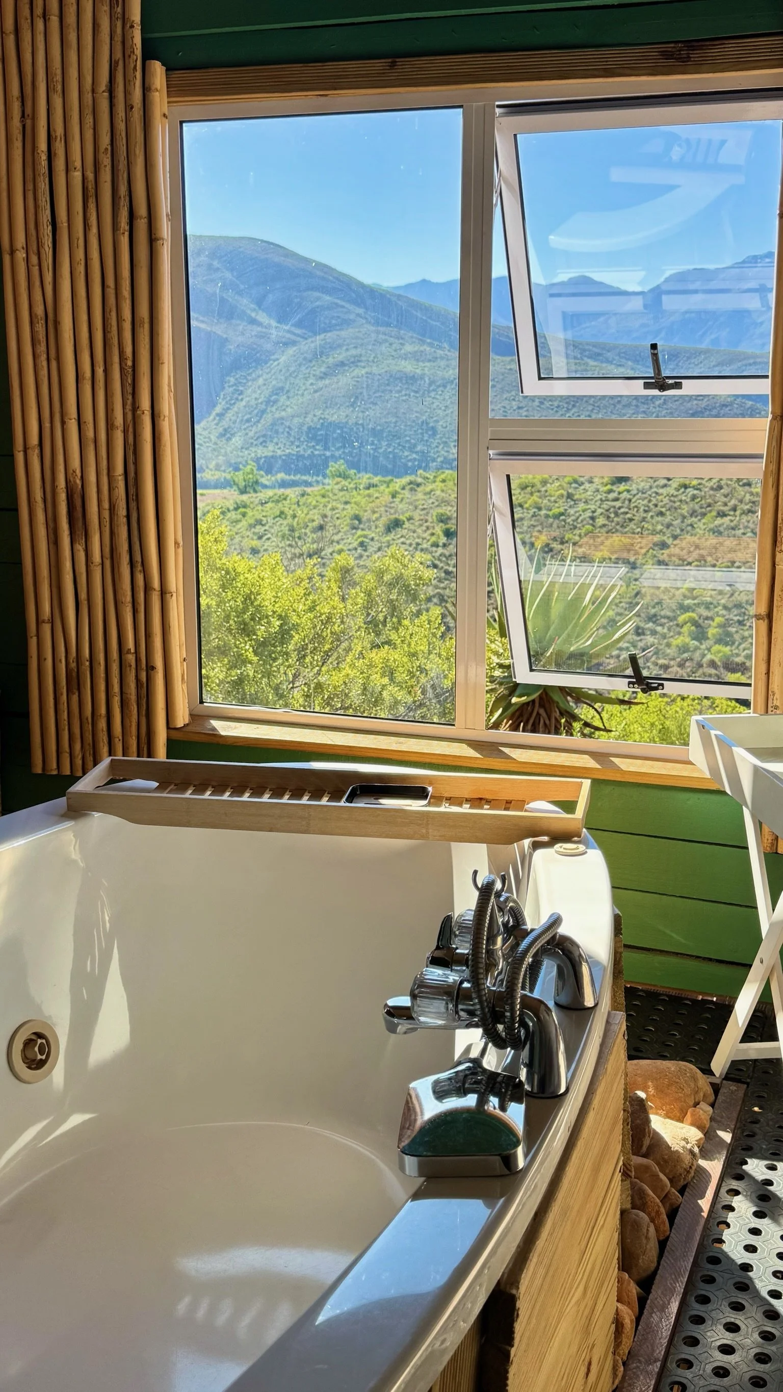 View from inside a room with bamboo walls and a large window showing mountains and greenery outside. An open window is visible, allowing fresh air inside. There is a bathtub with a wooden tray and a faucet, small rocks lining the side, and a white chair next to the tub.