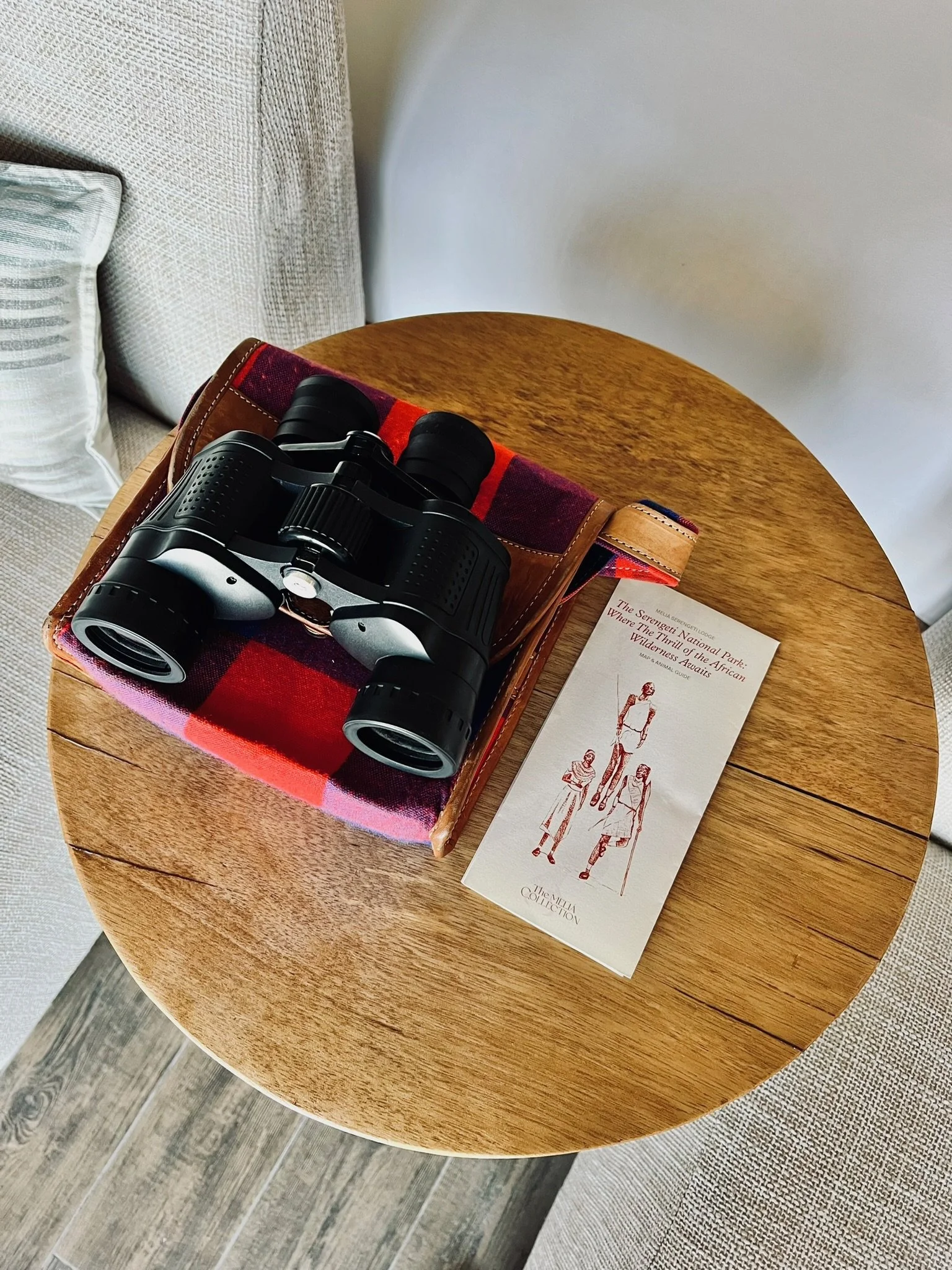 A pair of black binoculars resting on a red and black plaid cloth on a round wooden table, with a brochure featuring fashion sketches and text, next to a beige cushion with a subtle pattern.