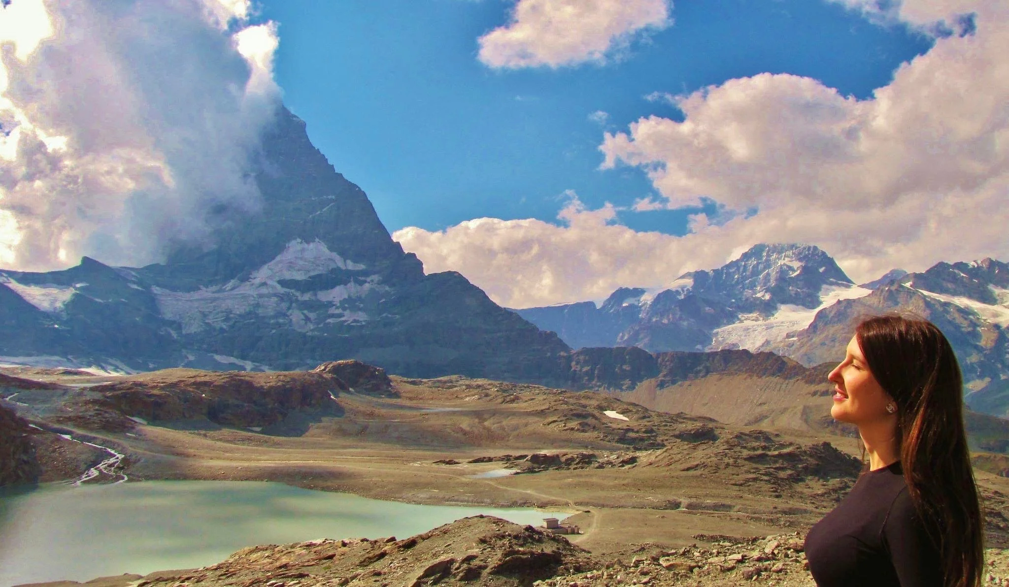 A woman with long brown hair and earrings smiling in profile outdoors in front of a lake with mountains, snow-capped peaks, clouds, and a blue sky.