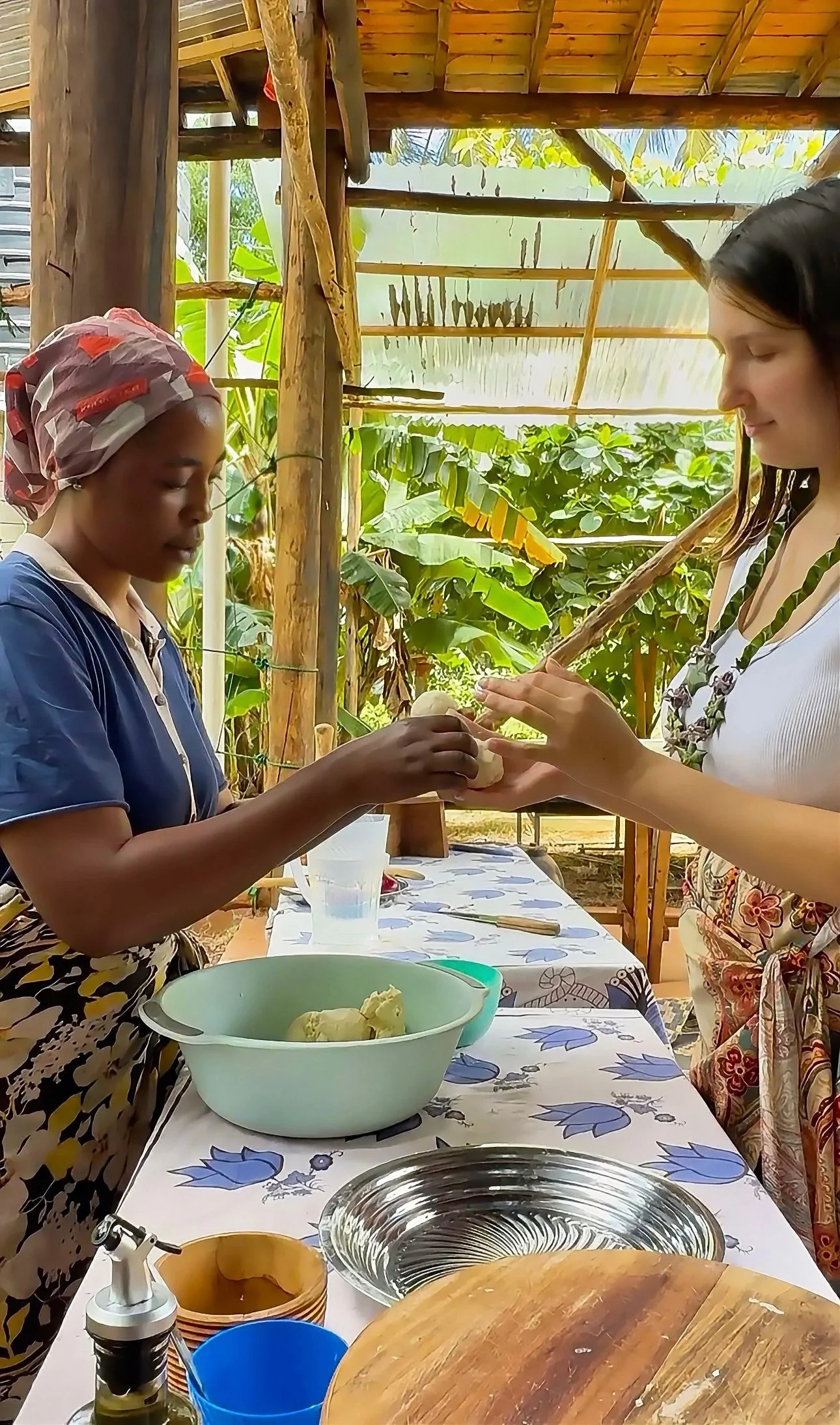 Two women are working together to prepare dough in an outdoor kitchen with lush green plants in the background. One woman is handing dough to the other woman.