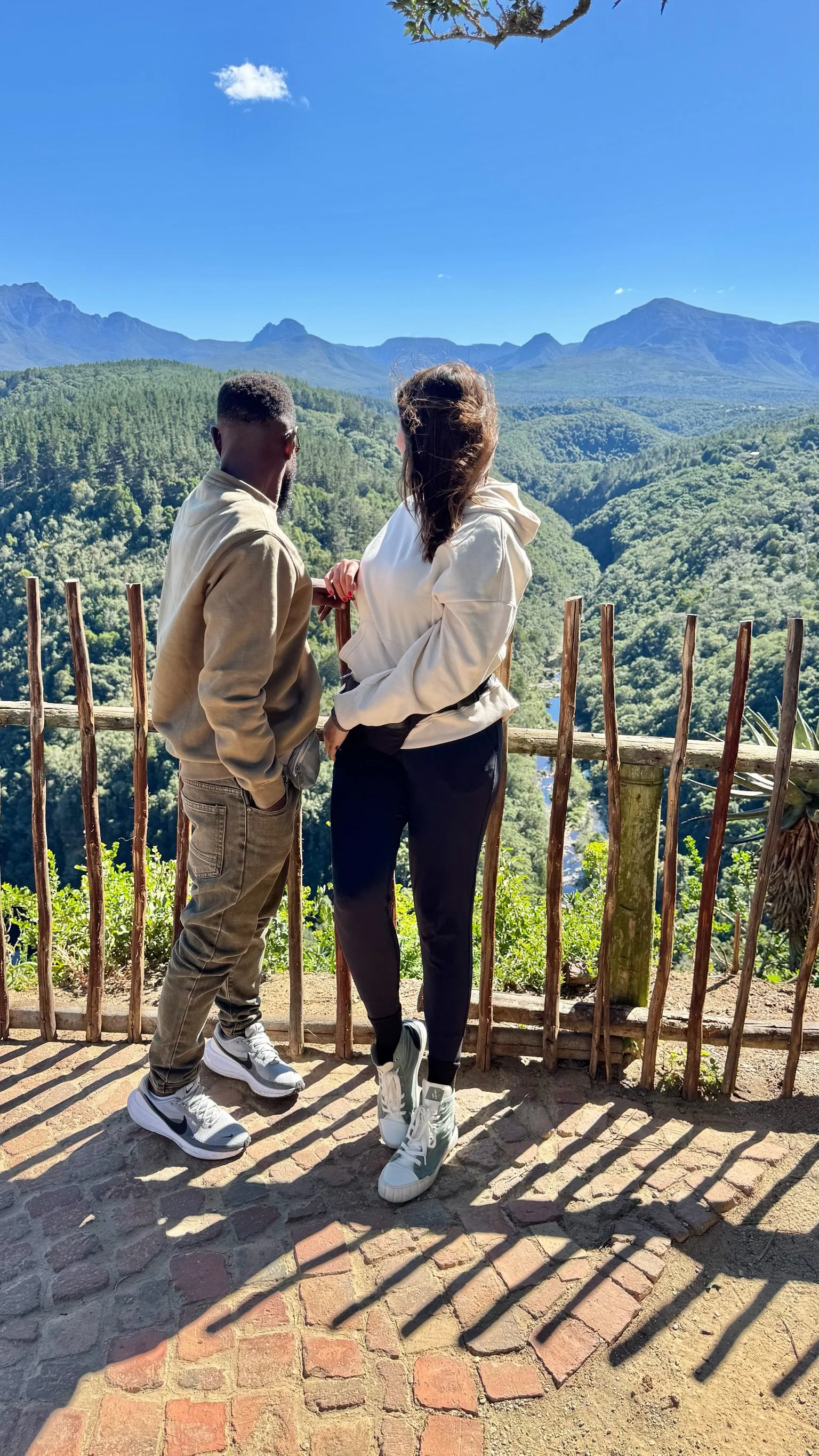 A man and woman stand on a viewing platform overlooking a lush green valley and mountain range, with clear blue skies above.