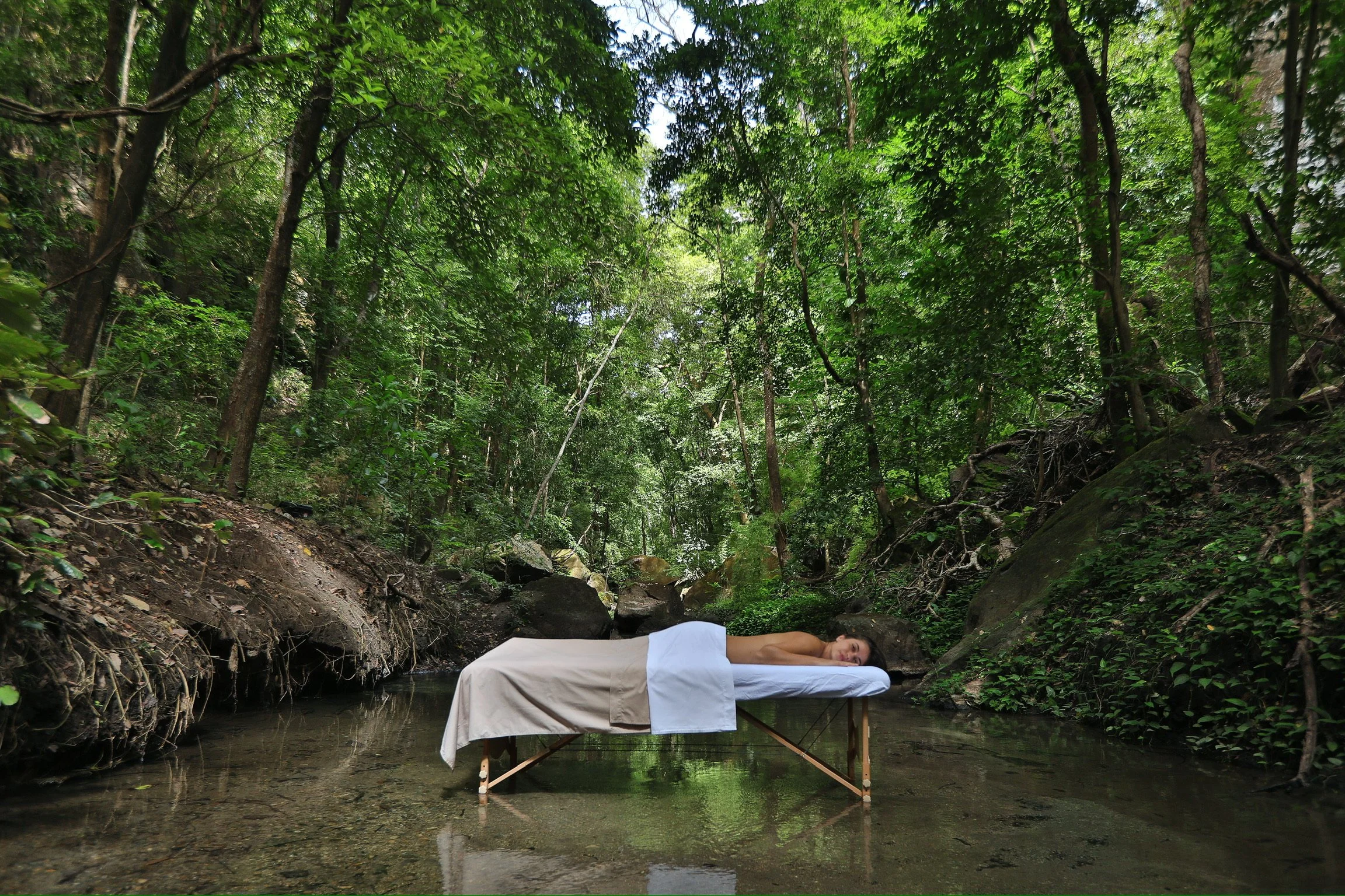 Person lying on a table covered with a white cloth in a forest creek surrounded by trees and greenery.