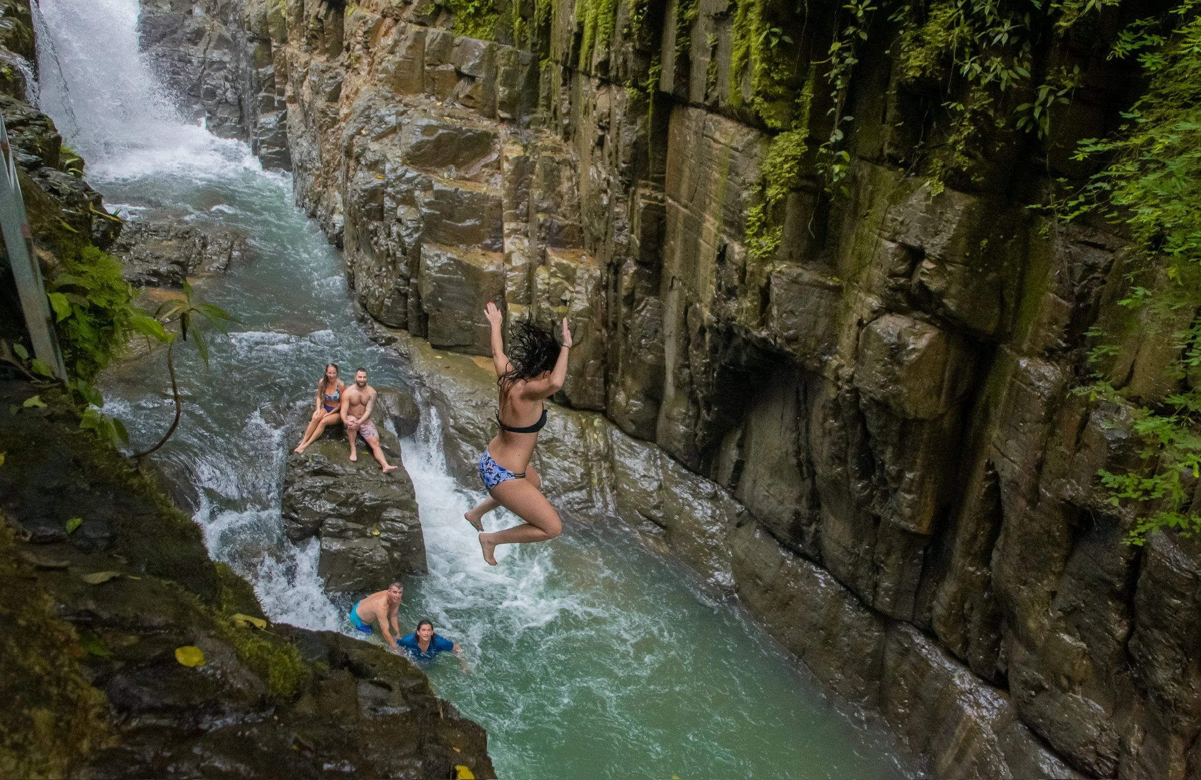 Two women and three men enjoying swimming and jumping in a rocky canyon with water and a small waterfall, surrounded by lush green foliage.