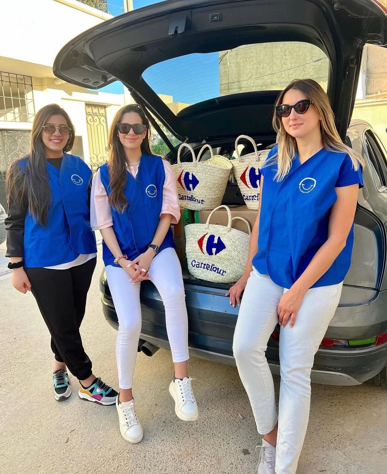 Three women standing beside an open car trunk filled with Carrefour shopping bags. They are outdoors, smiling, and wearing blue Carrefour vests and sunglasses.