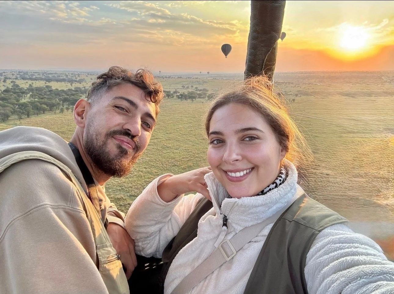 A smiling man and woman taking a selfie during a hot air balloon ride at sunset over a grassy landscape with trees and other hot air balloons visible in the background.