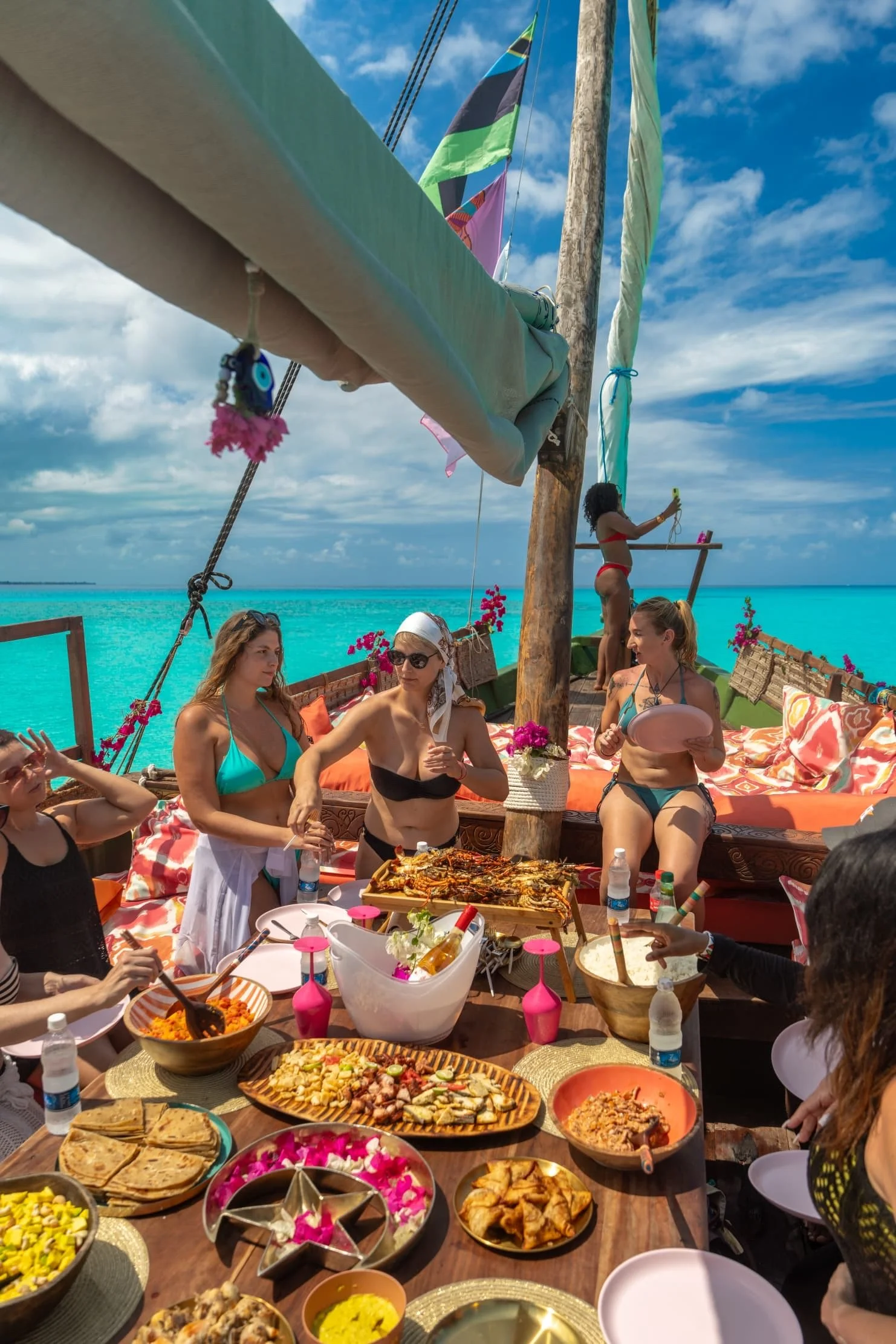 People gathered around a table of various dishes, outdoor setting with bright blue sky, women in swimsuits, some wearing hats, decorations, and a woman taking a selfie. The scene appears to be at a beachside or tropical location.