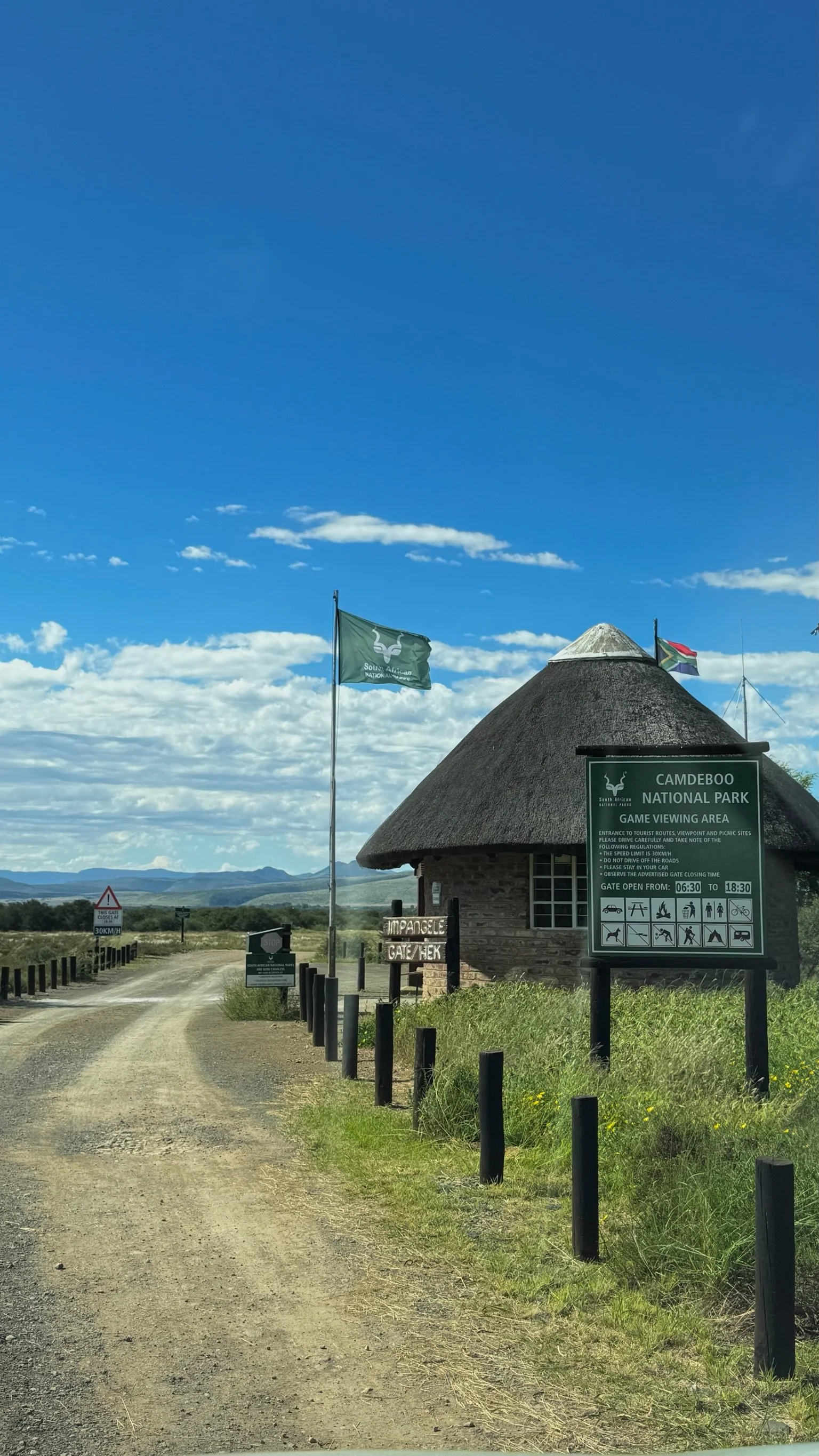 Entrance to Camdeboo National Park with a thatched building, signs, flags, and a dirt road leading into the park under a partly cloudy blue sky.