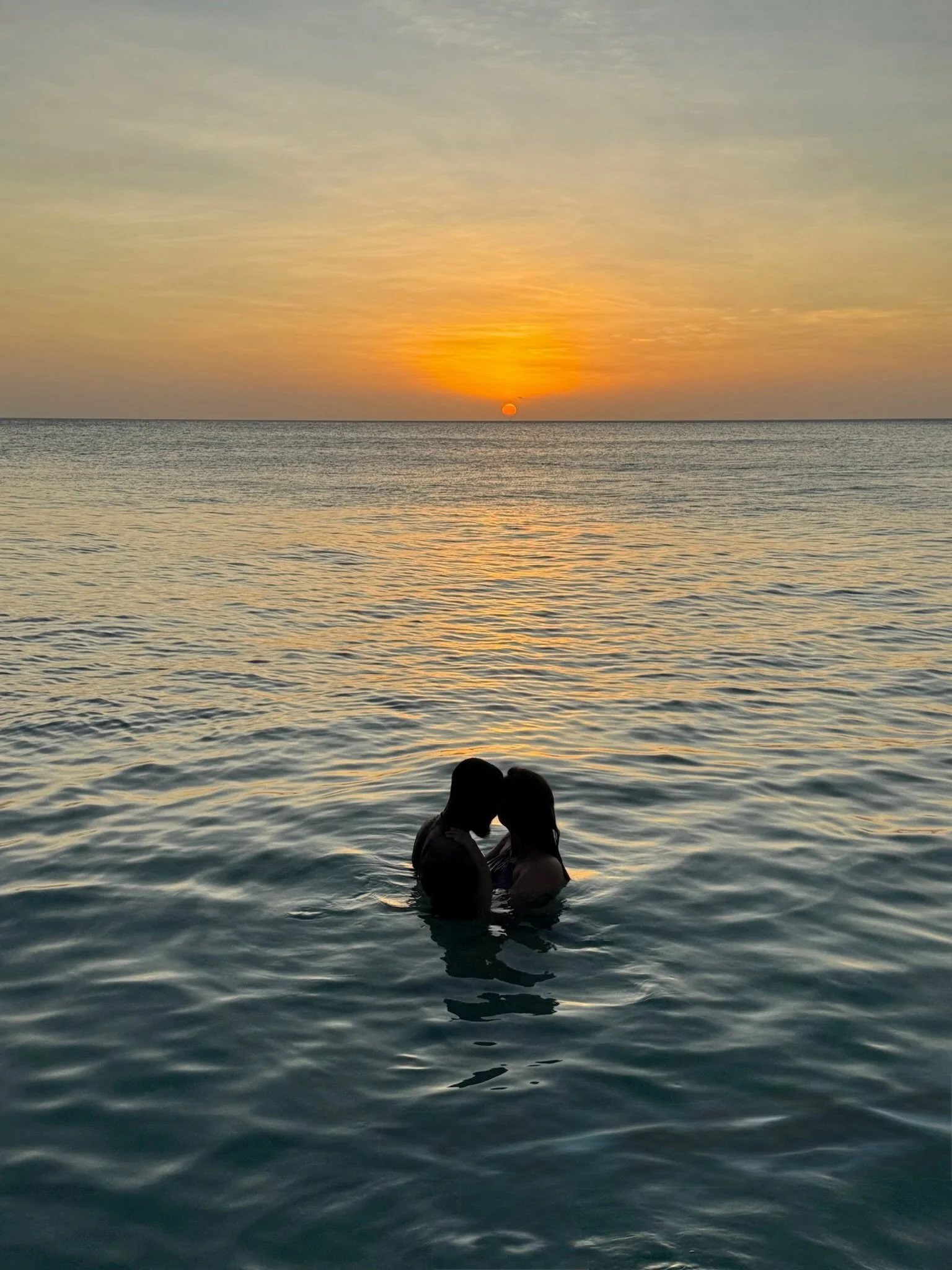 A couple is sharing a kiss in the ocean with the sun setting on the horizon, creating a romantic scene with warm colors in the sky.