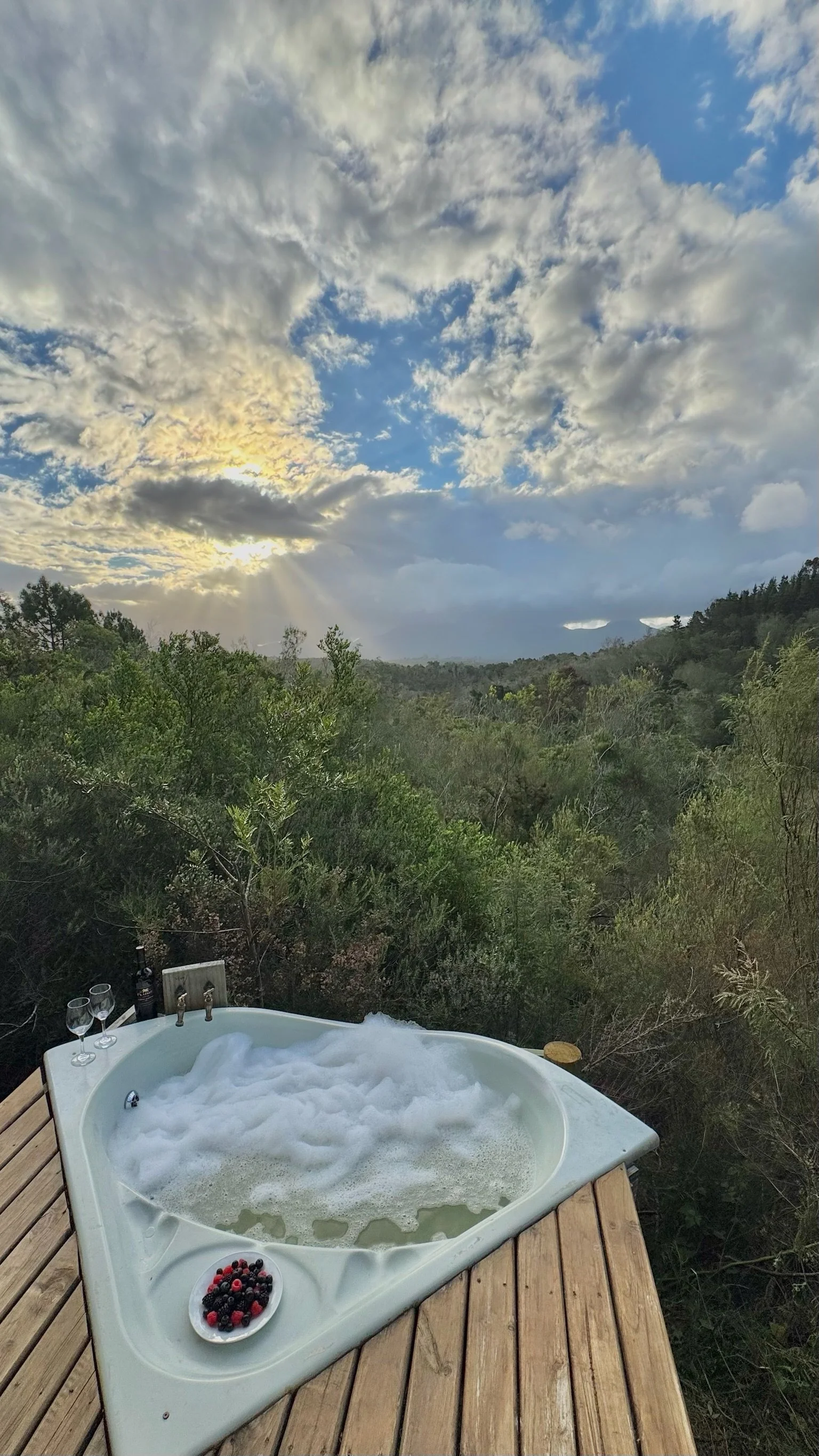 An outdoor hot tub filled with bubbles on a wooden deck, overlooking a lush green hillside with a cloudy sky and sunlight rays.
