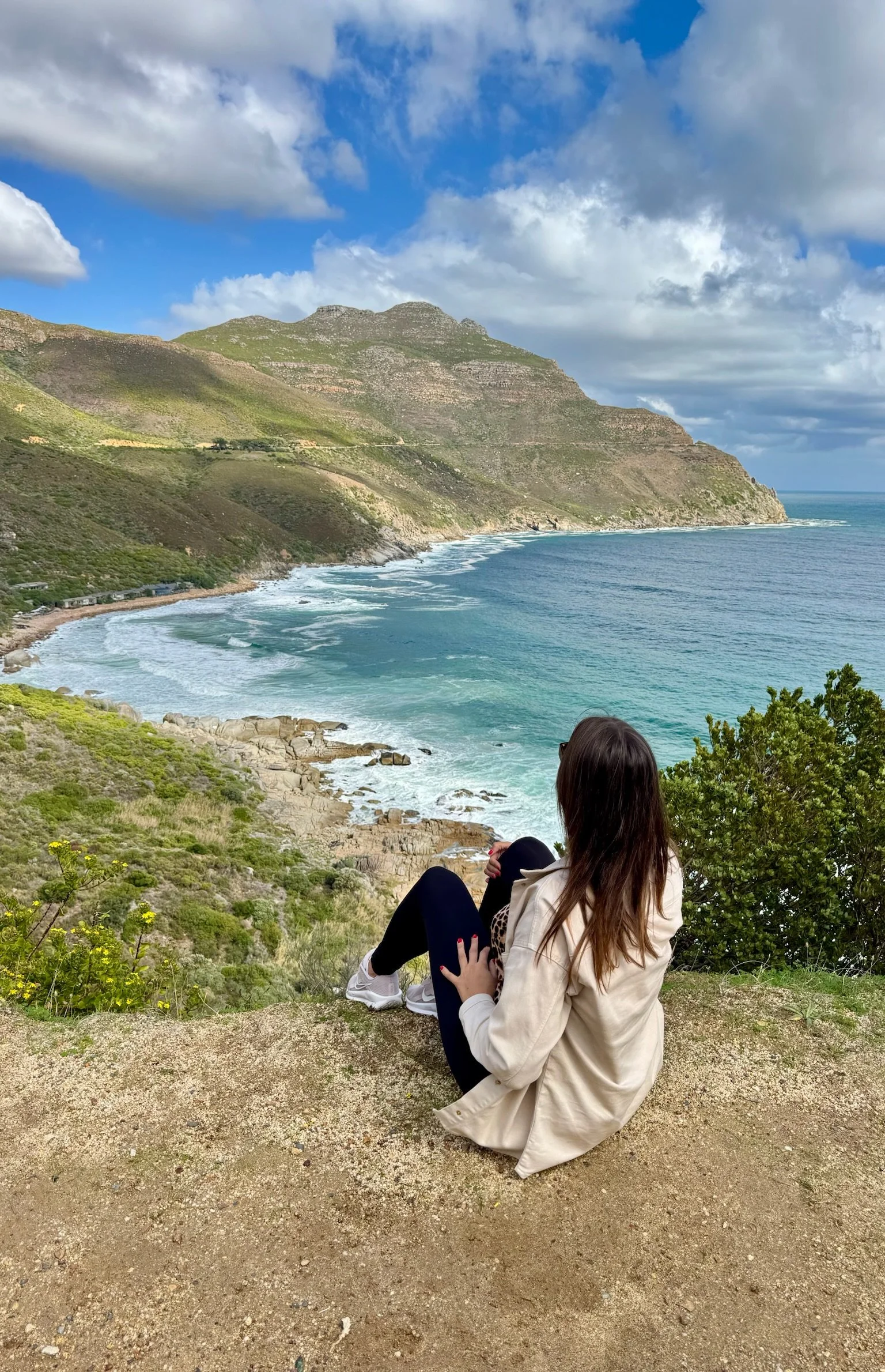 A woman with long brown hair sitting on a grassy hill, overlooking the ocean with waves crashing along the shore. She is wearing black leggings, white sneakers, a light beige coat, and sunglasses, and appears to be relaxing and enjoying the scenic view of mountains and cloudy sky.