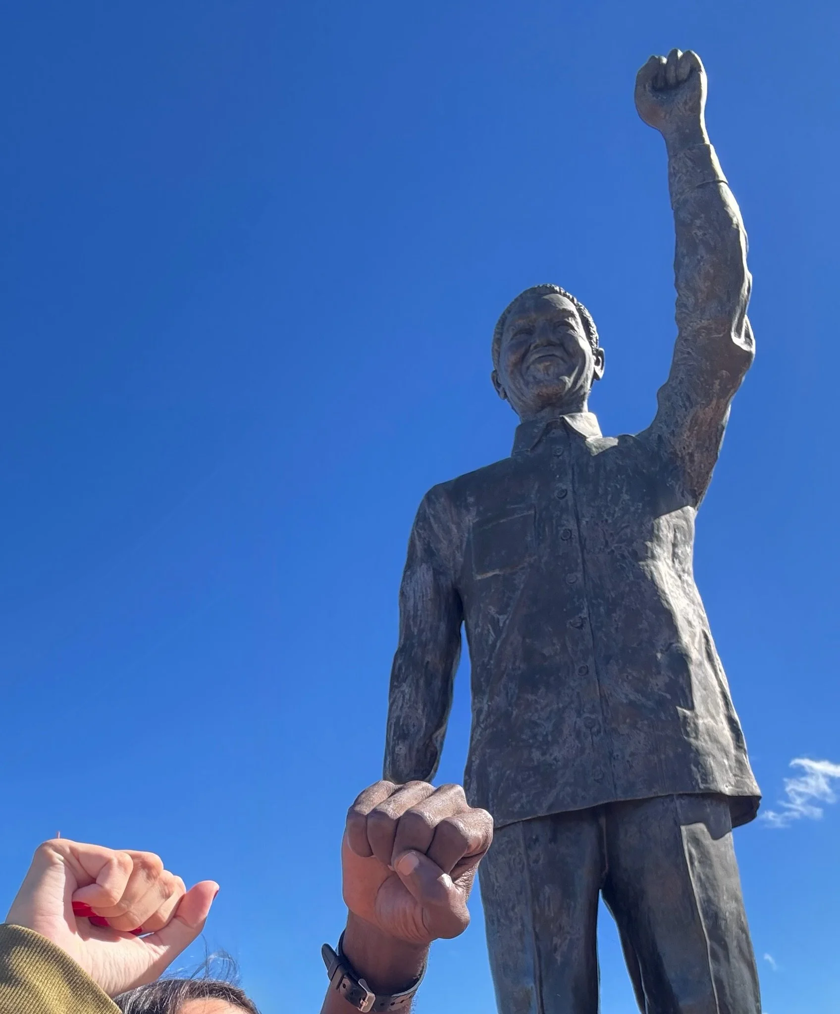 A person clenches their fist and raises their arm, interacting with a large statue of a smiling man in a suit raising his hand, against a clear blue sky.