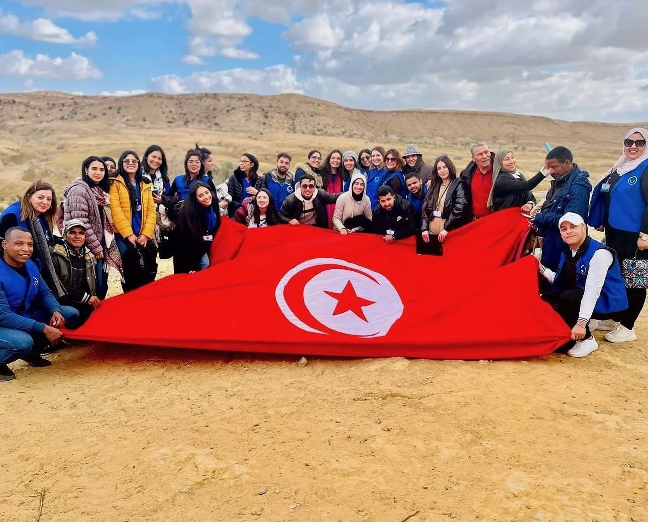 Group of people outdoors holding a large Tunisian flag with a red background, white circle, crescent moon, and star, in a desert landscape with hills and clouds in the sky.