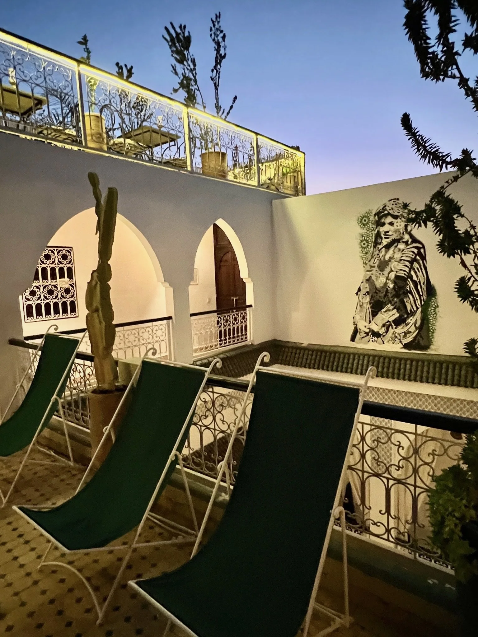 Outdoor patio area at dusk with lounge chairs, potted plants, artwork of a person in black and white, and a decorative iron balcony railing.