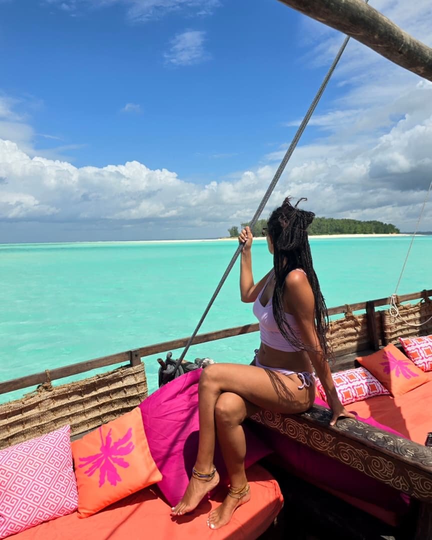 Woman in white swimwear sitting on a boat with coral cushions, holding a rope, overlooking turquoise water and a cloudy sky.