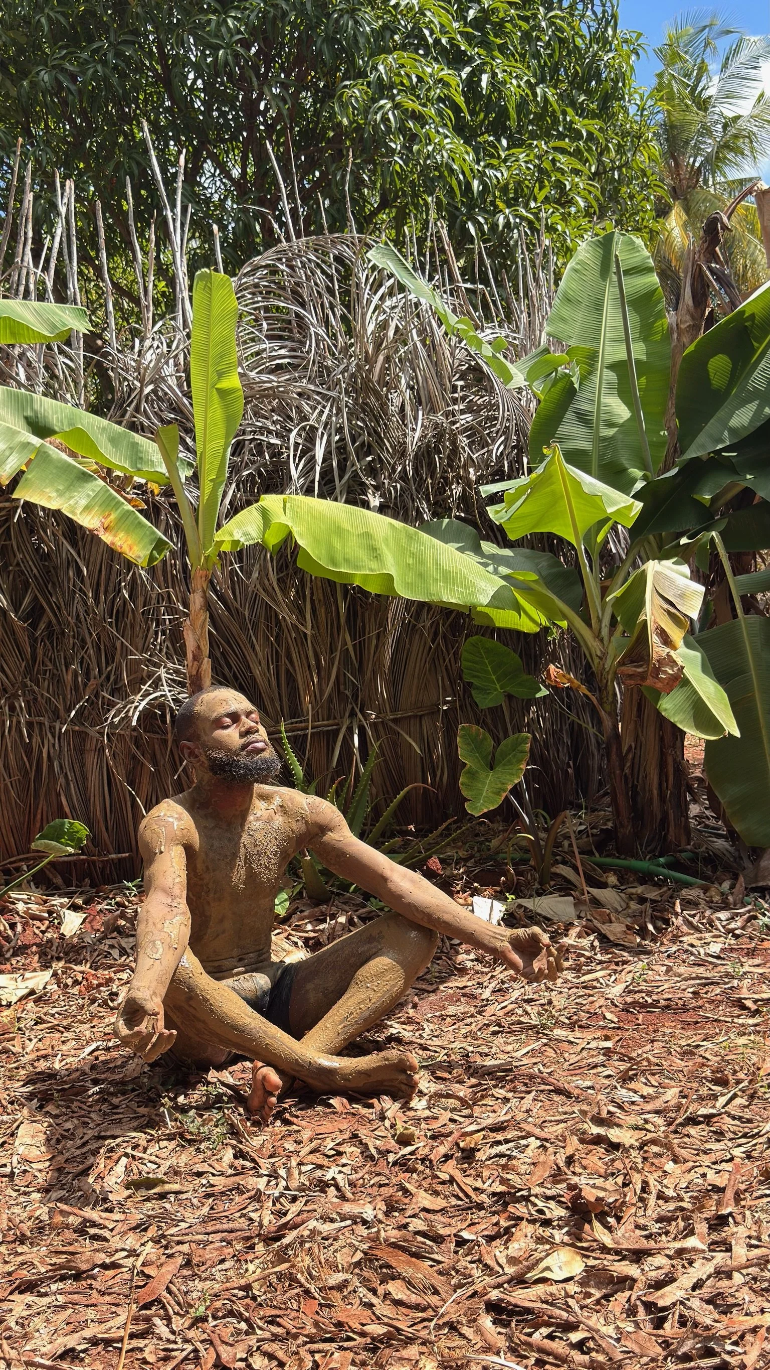 A man with mud on his body practicing yoga or meditation outdoors among lush green jungle plants under a blue sky.