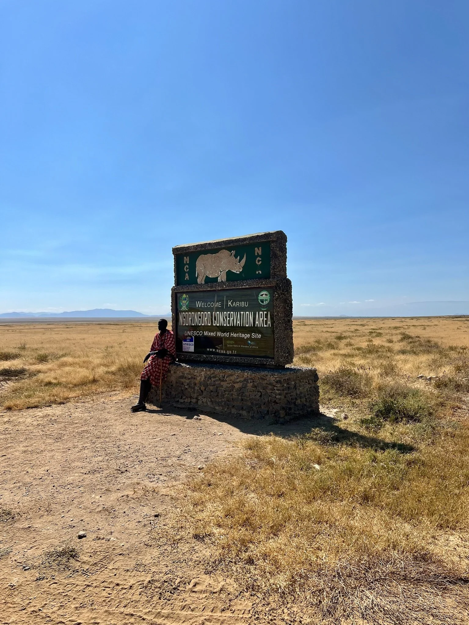 A large signboard at Ngorongoro Conservation Area in Tanzania with a rhinoceros illustration and welcoming text, situated in a vast open landscape with dry grass, a person sitting next to the sign, mountains in the distance, and a clear blue sky.