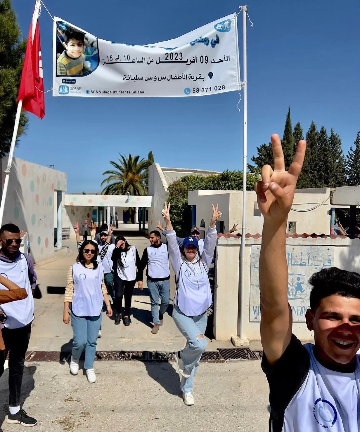 Group of people walking outside a building, some making peace signs. They are wearing white vests with logos, and there is a banner overhead with Arabic text and a photo of a young boy. The background shows a clear blue sky, trees, and a white building.