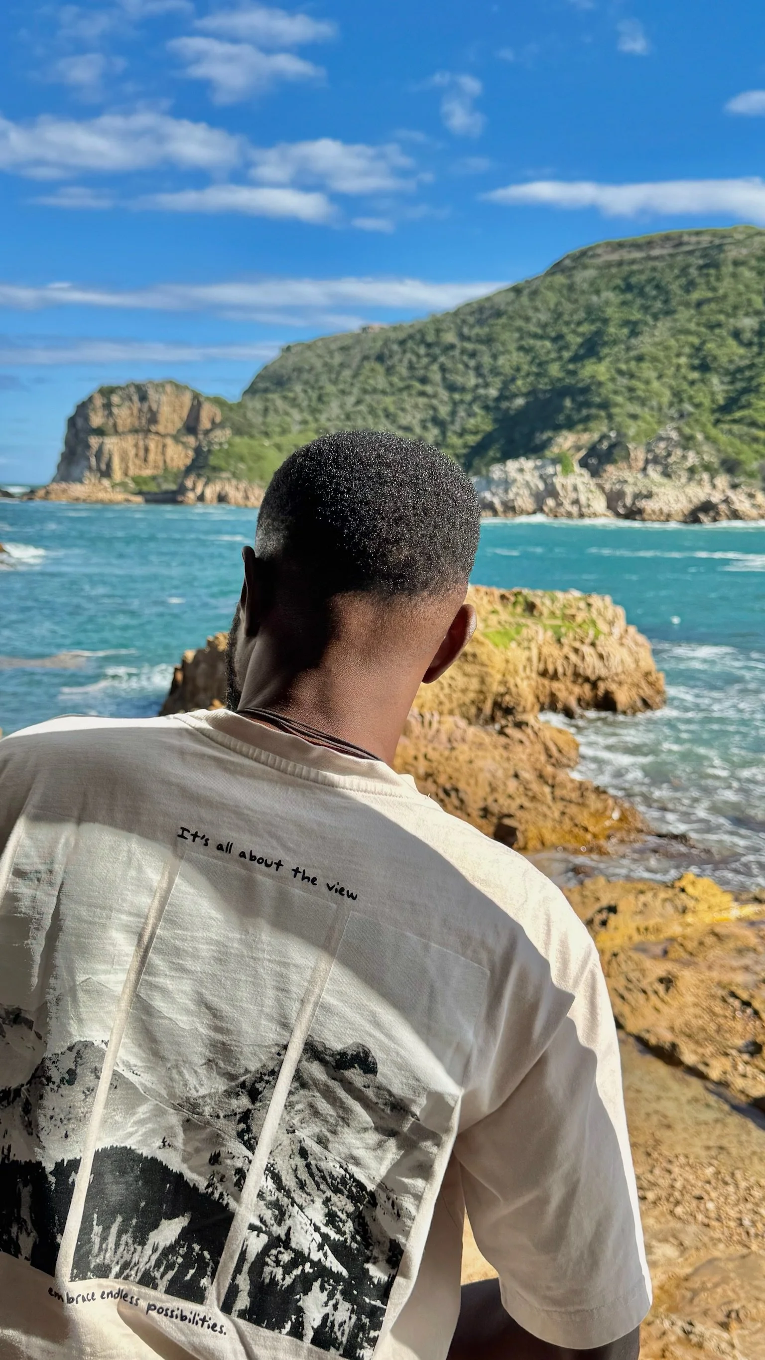 A person with short hair facing away, wearing a white shirt with mountain and nature print, looking at a coastal landscape with rocky shore, blue ocean, green hills, and a partly cloudy sky.