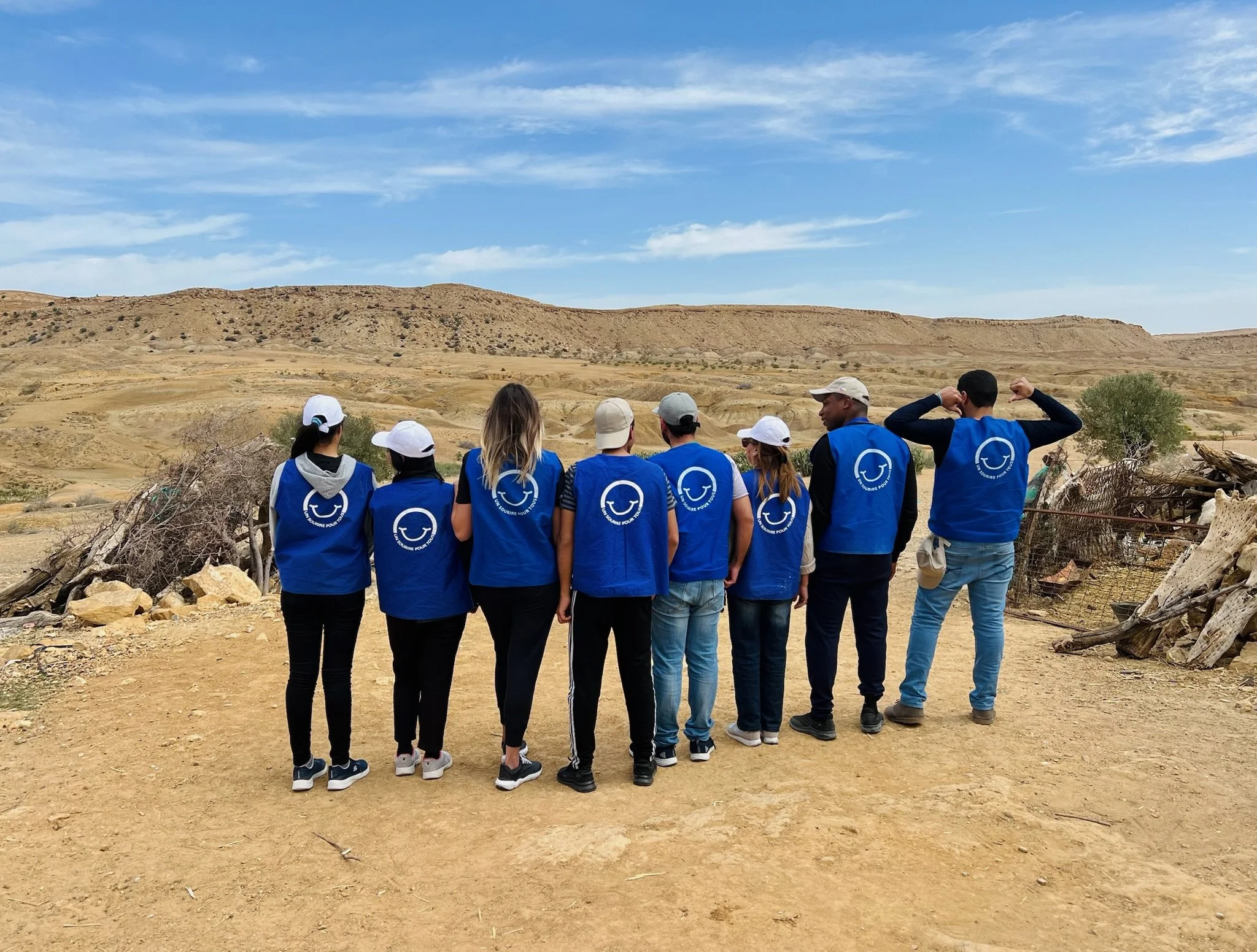 A group of people wearing blue vests and white caps standing in a desert landscape, looking towards distant hills under a blue sky with some clouds.