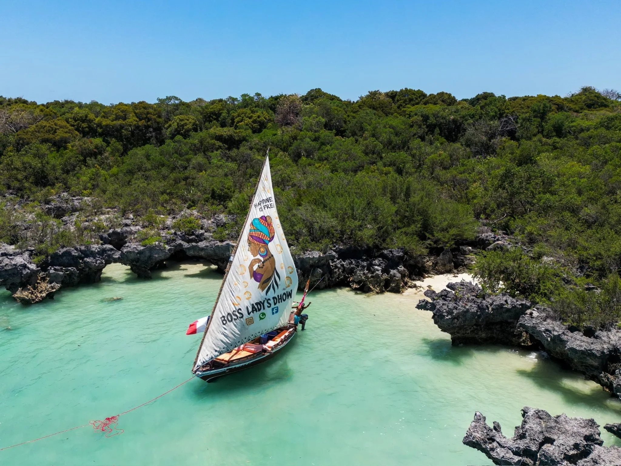 A boat with a sail featuring a colorful illustration of a woman with a headwrap and the text 'Boss Lady's Dhow' on a turquoise river, surrounded by rocky banks and dense green trees under a blue sky.