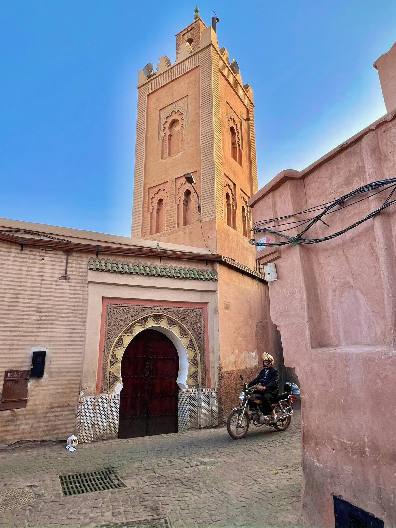 A tall, ornate tower with a clock and decorative windows, made of reddish-brown bricks, set against a clear blue sky. The street below shows a man riding a motorcycle and some architecture with intricate designs around a gated entrance.