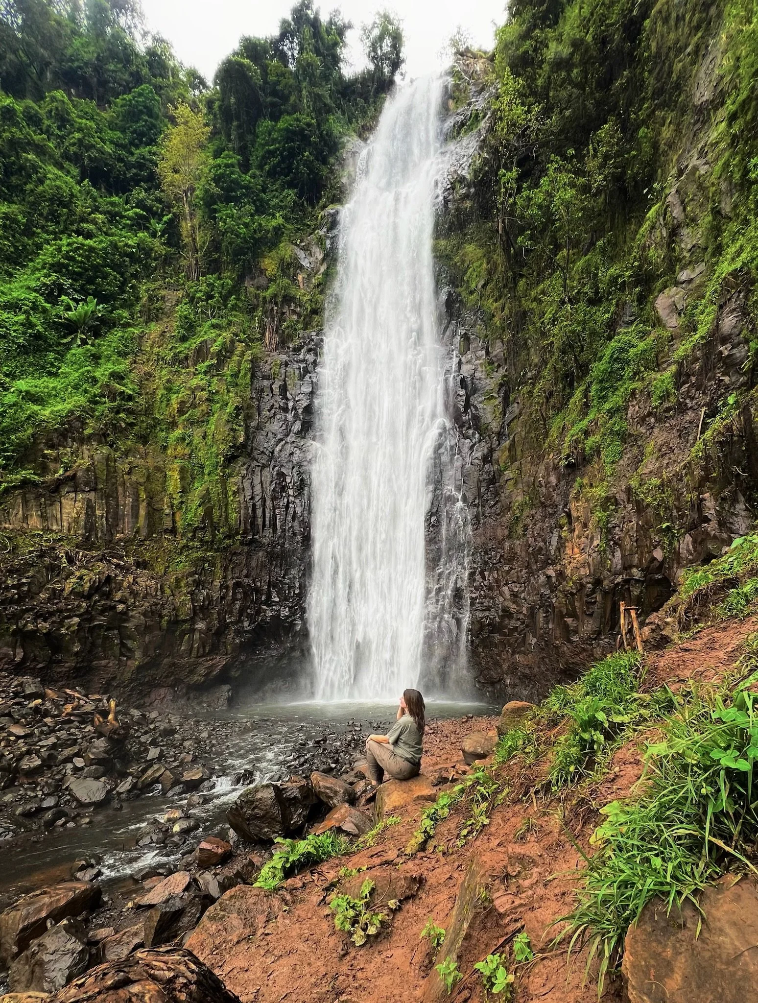 A person sitting on a rock by a river at the base of a tall waterfall surrounded by lush green vegetation and rocks.