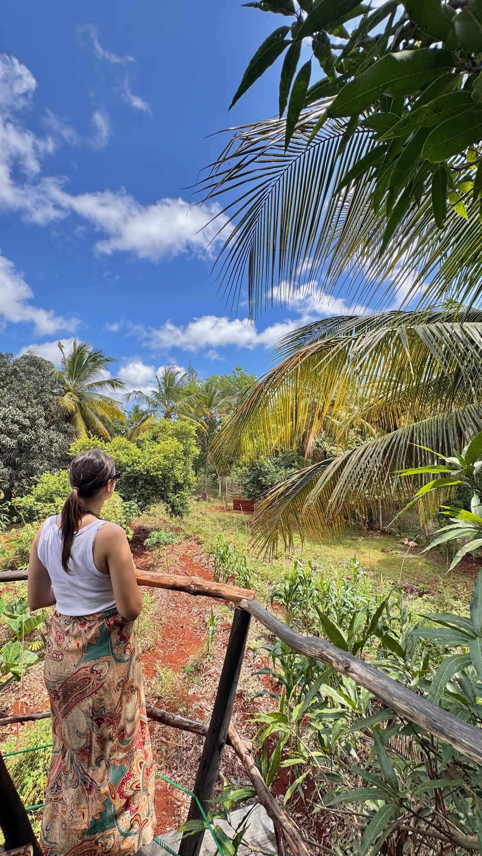 A woman with brown hair tied back, wearing sunglasses, a white sleeveless top, and a patterned skirt, standing on a wooden bridge overlooking a lush garden with green plants and trees, under a bright blue sky with scattered white clouds.