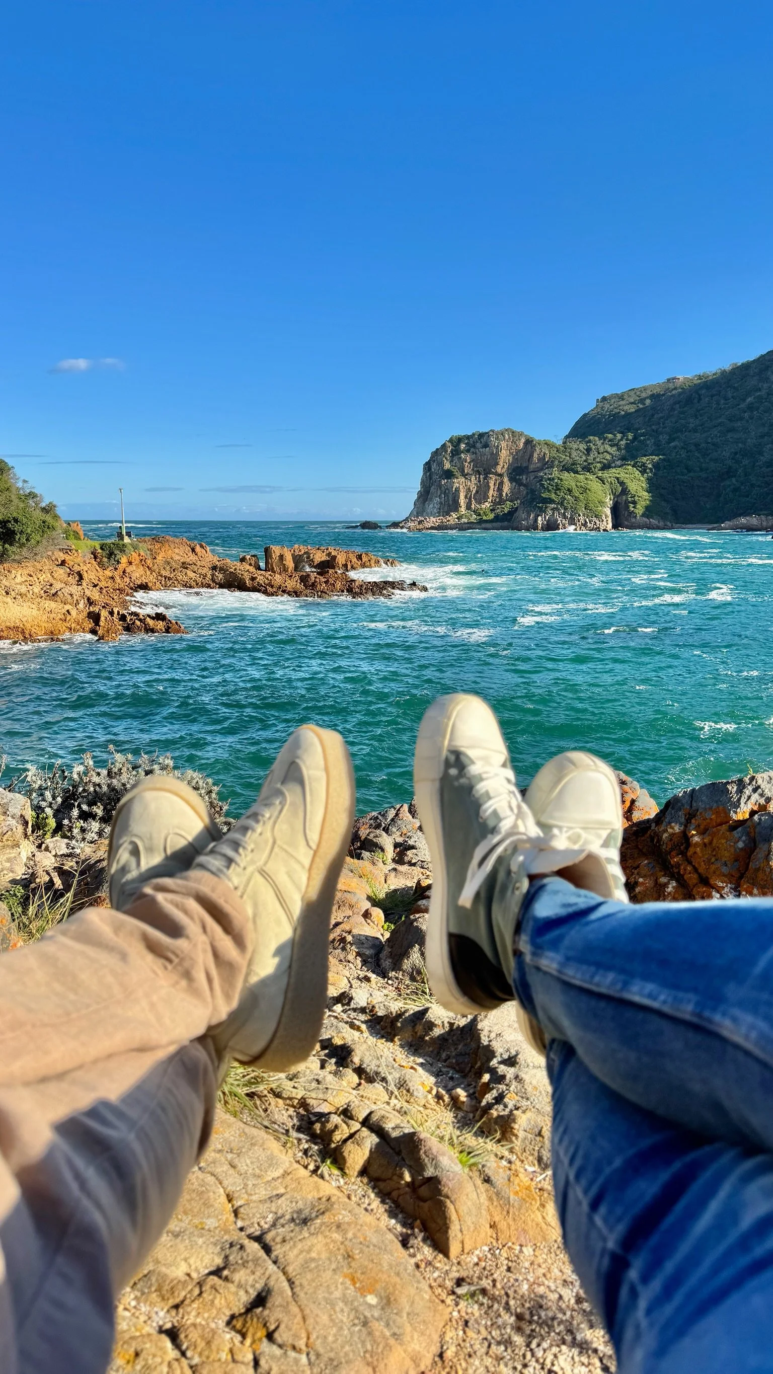 Two people with sneakers visible, sitting on rocky shore, facing ocean with waves, cliffs, and clear blue sky.