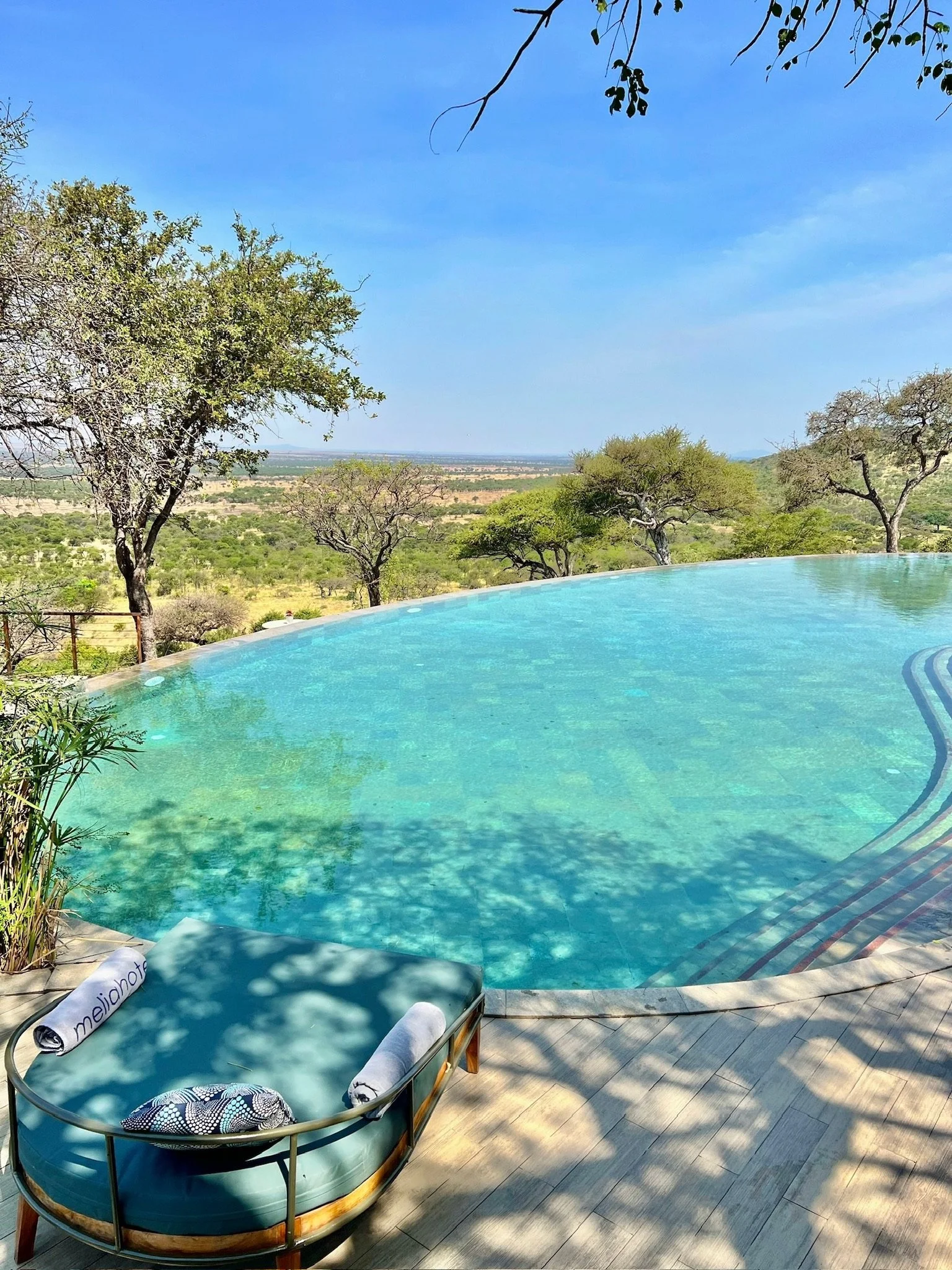 A swimming pool with a curved edge, surrounded by a wooden deck. Two lounge chairs with rolled towels and a decorative cushion are positioned in the shade. There are trees and a scenic landscape in the background with a clear blue sky.