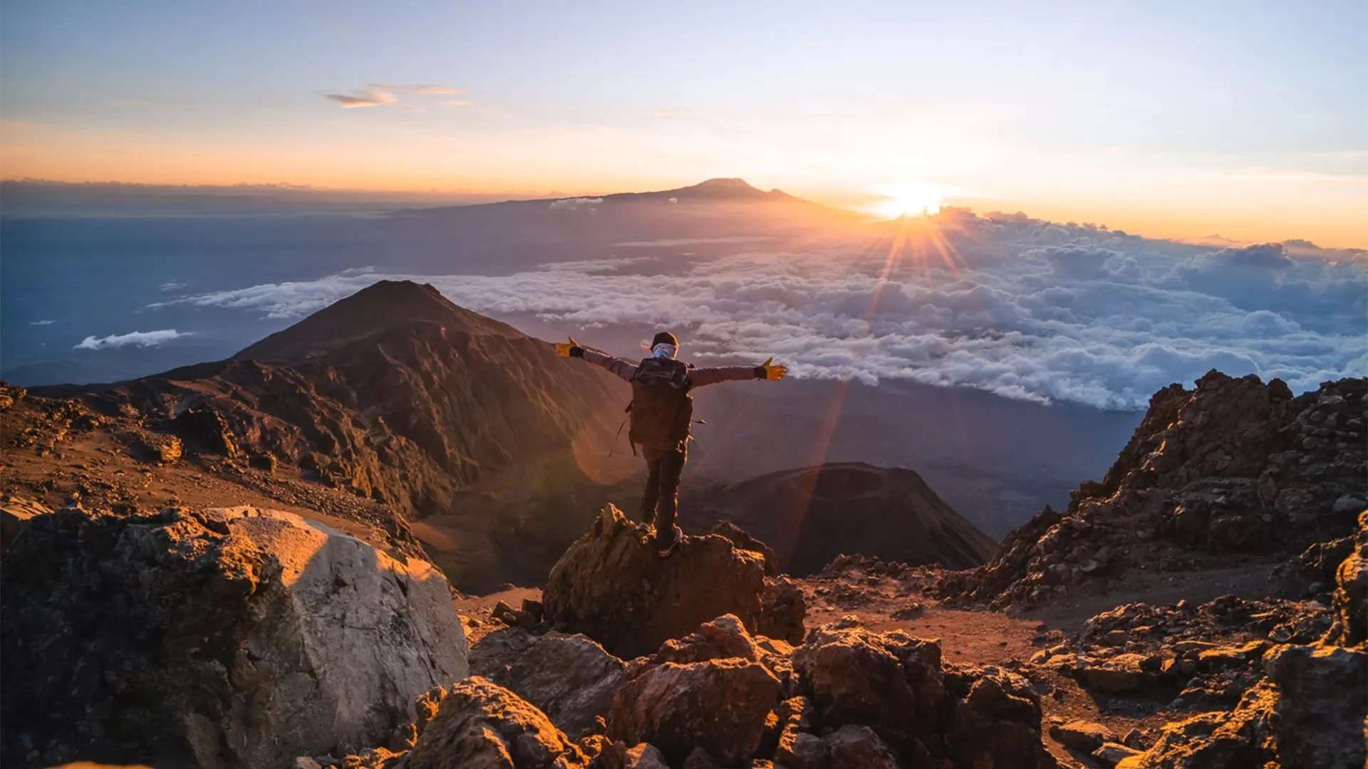 A person standing on a rocky mountain summit with arms outstretched, facing a sunrise above clouds and distant mountains, including a volcano.
