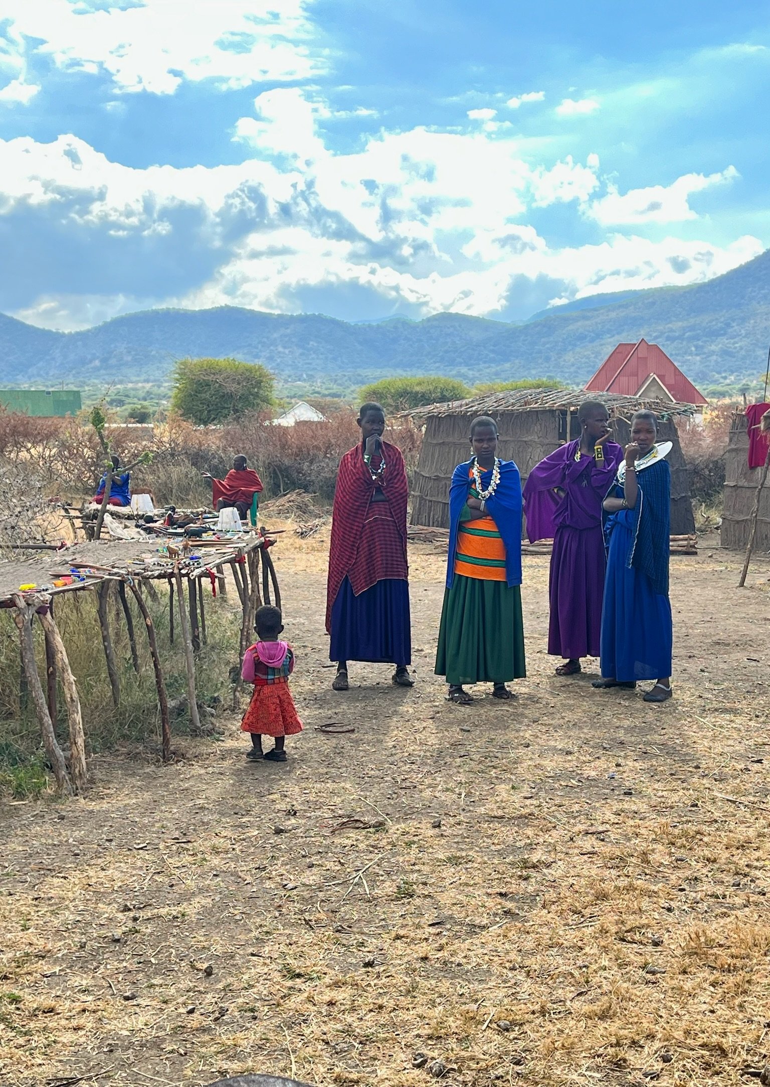 Four women in traditional colorful clothing and jewelry, and two children in rural outdoor setting with adobe houses, trees, mountains, and partly cloudy sky in the background.