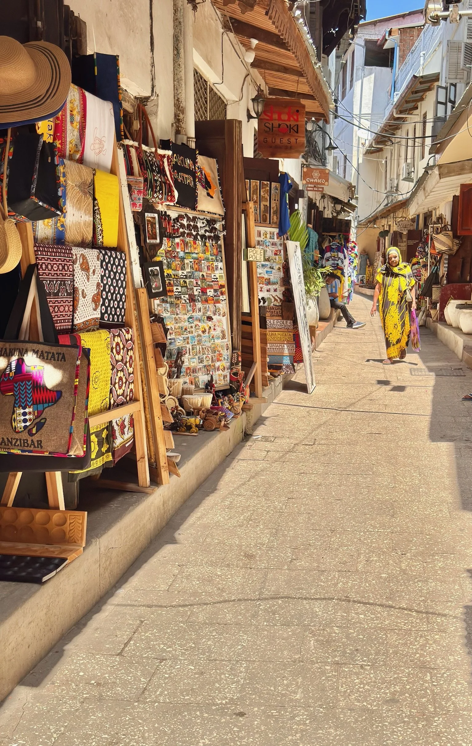 A street market with various souvenirs and clothing displays on the left side, including hats, bags, and small decorative items. A woman dressed in yellow with a head covering is walking down the street, and a person sits in the background. The street is narrow with buildings on both sides, and signs hang overhead.