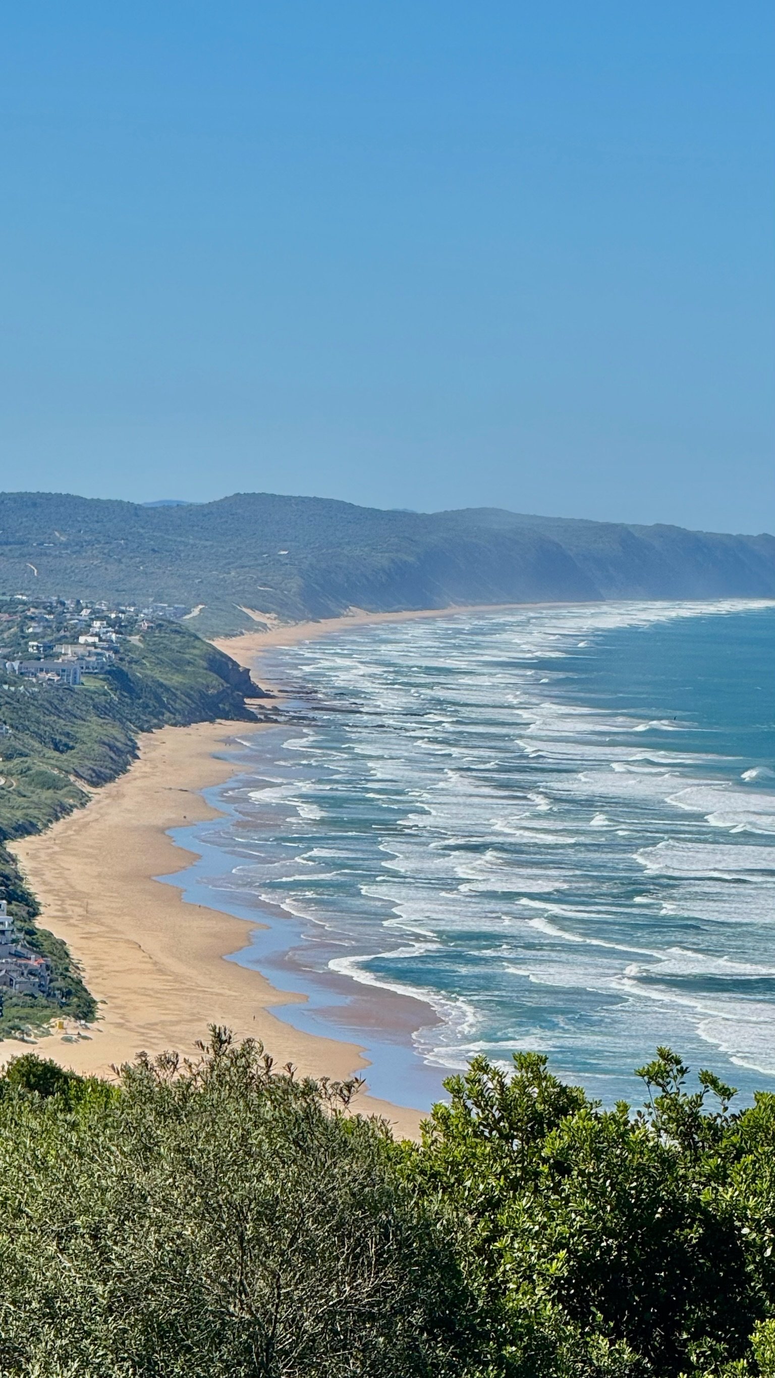 A scenic view of a coastline with sandy beach, ocean waves, green hills, and a clear blue sky.