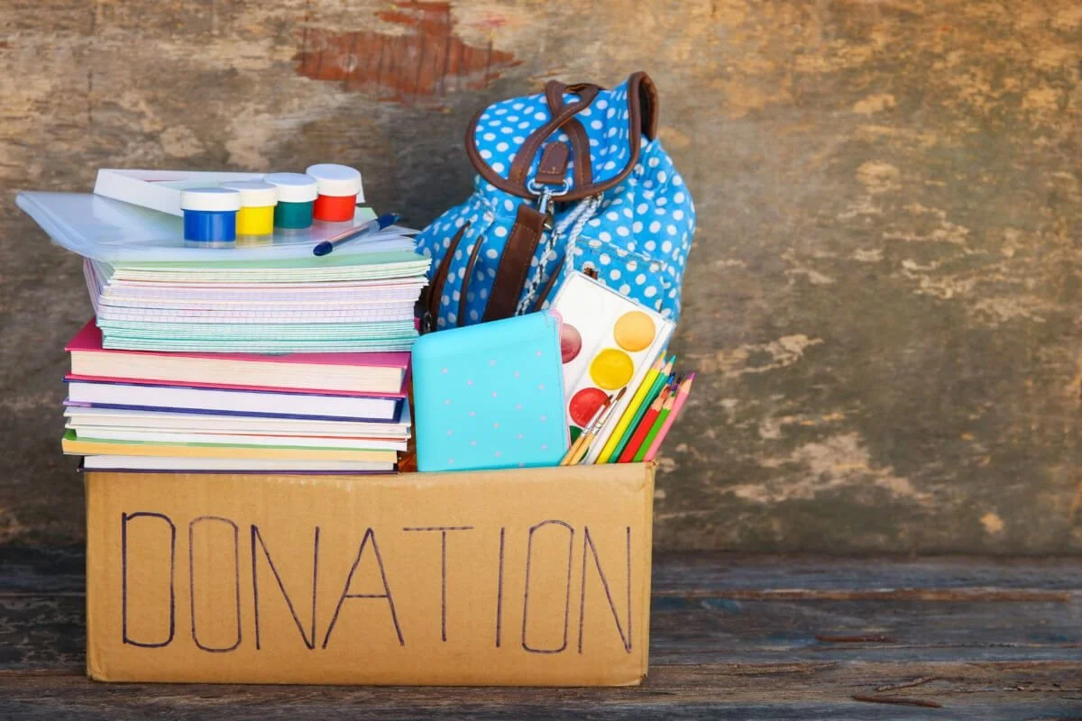 Open cardboard box labeled 'Donation' filled with school supplies, including notebooks, paint sets, a backpack, colored pencils, and a watercolors set.