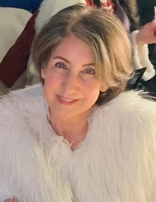 A smiling woman with short, light brown hair, wearing a white feathered or furry garment, at a social gathering or party.