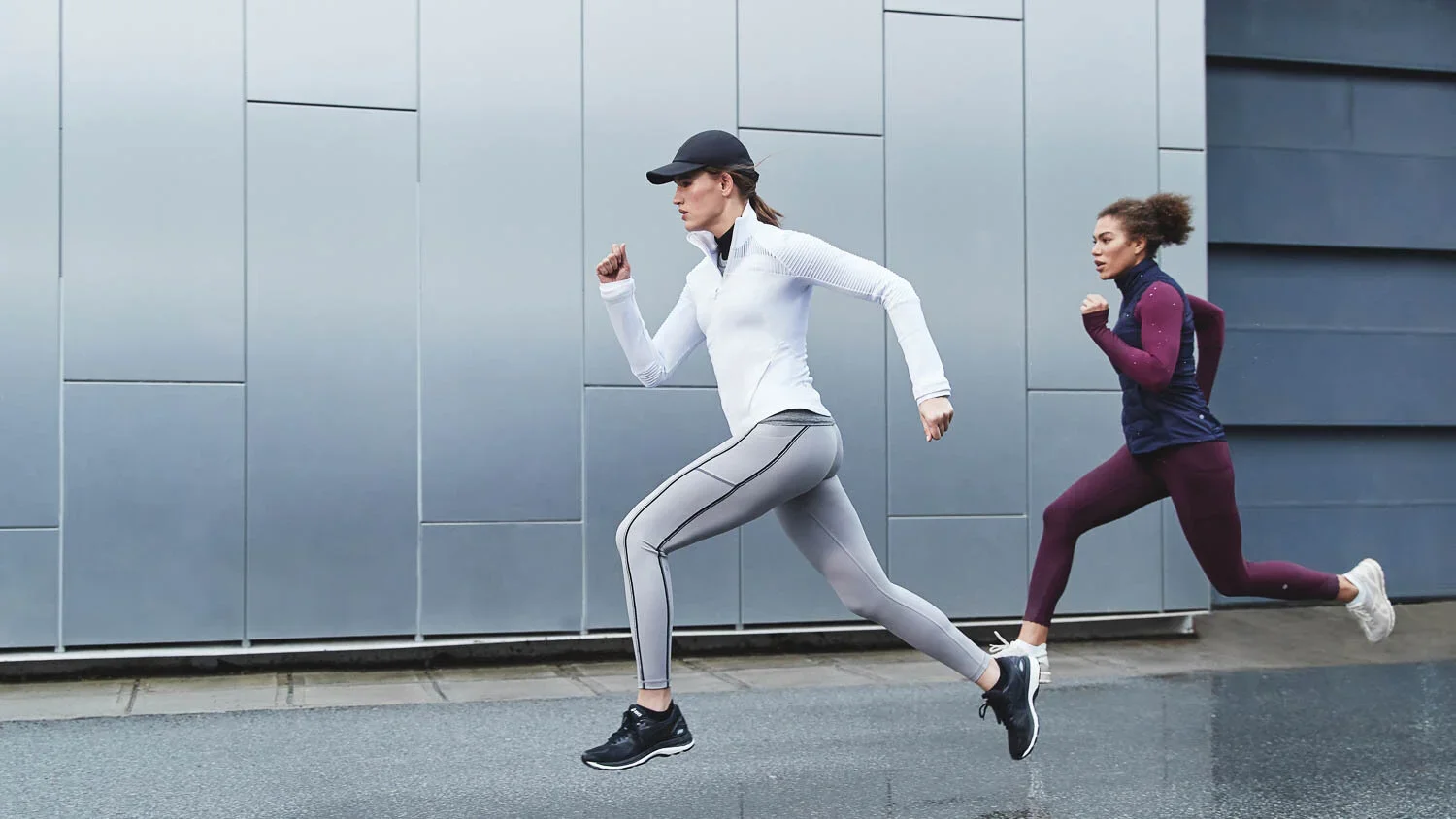 Two women running outdoors in workout clothes, one in front and one behind, against a modern gray building backdrop.