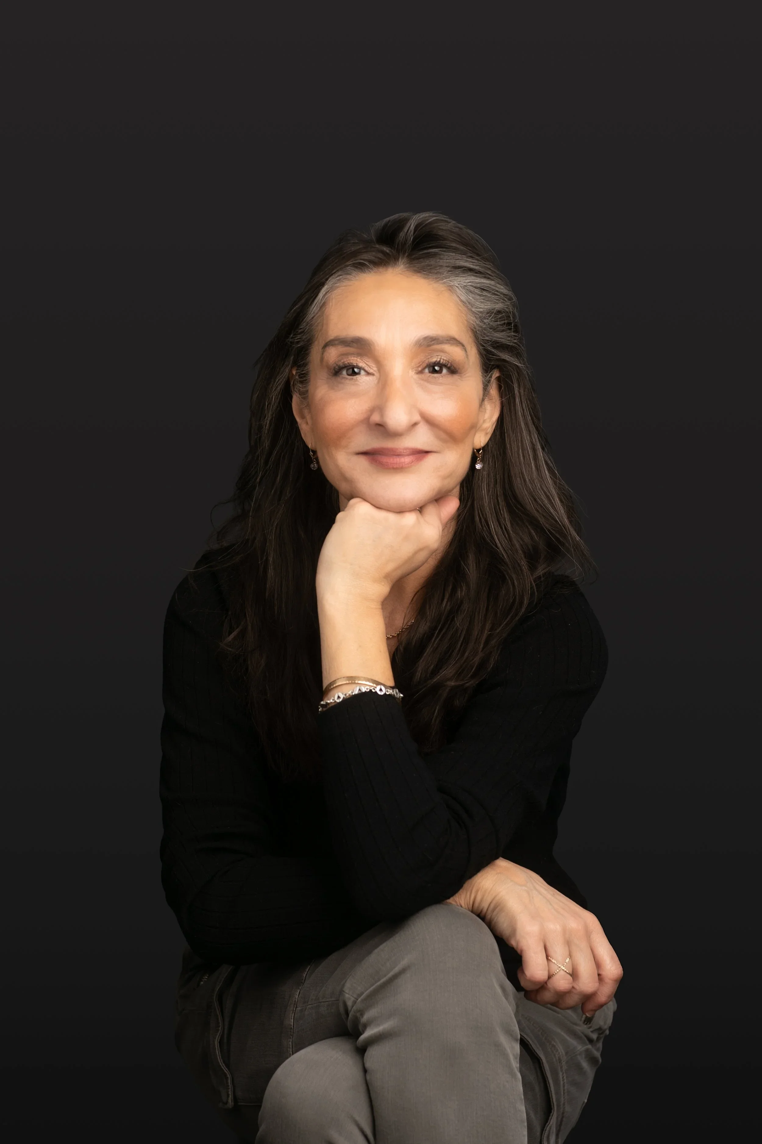 Portrait of a woman with long dark hair, smiling, resting her chin on her hand, against a dark background. She is wearing earrings, a watch, and a dark top.