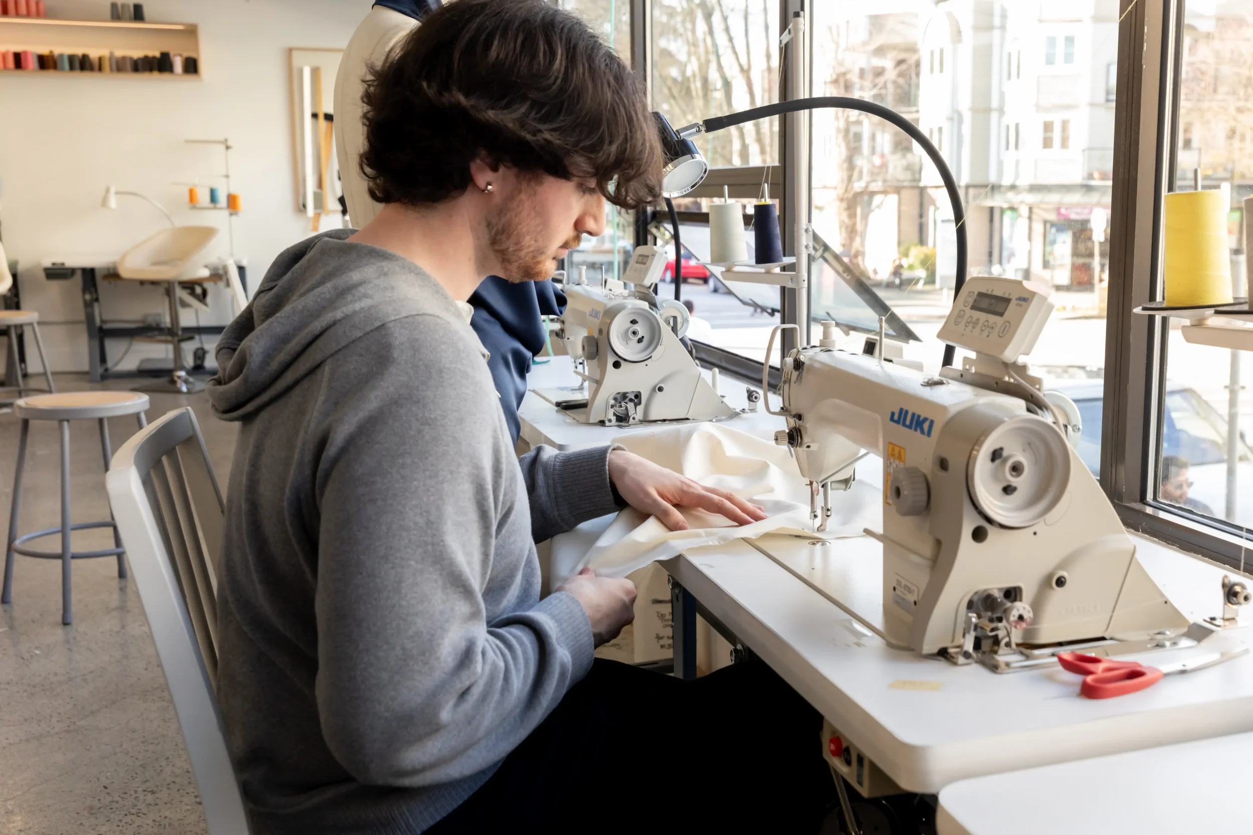 A person working in a sewing workshop using a Juki industrial sewing machine, with spools of thread and scissors on the table by a large window.
