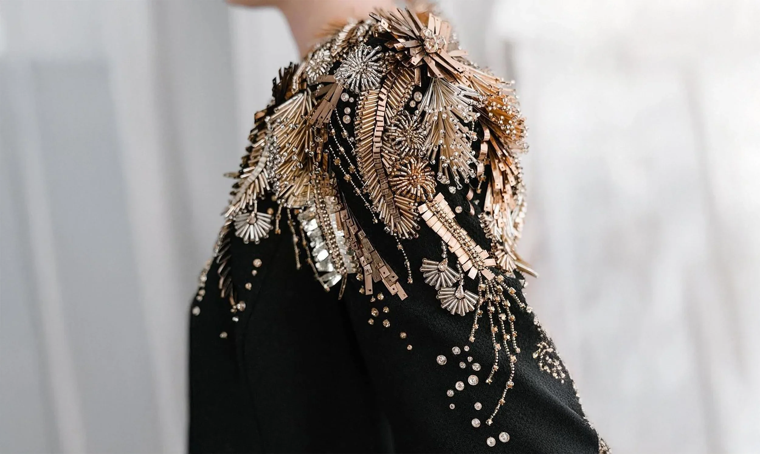 Close-up of a person's shoulder and side, wearing a black fabric garment embellished with numerous gold and silver metallic beads, sequins, and decorative elements arranged in floral and geometric patterns.