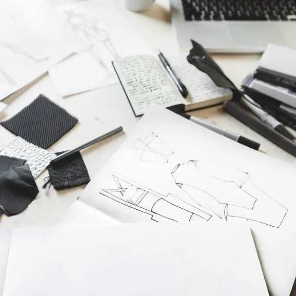 Desk with sketches of clothing designs, fabric swatches, pens, and an open notebook.