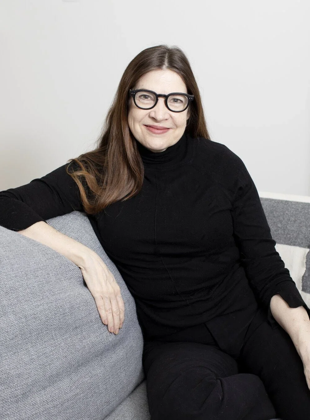 A woman with long brown hair, black glasses, and a black turtleneck, sitting on a gray couch against a plain white wall, smiling.