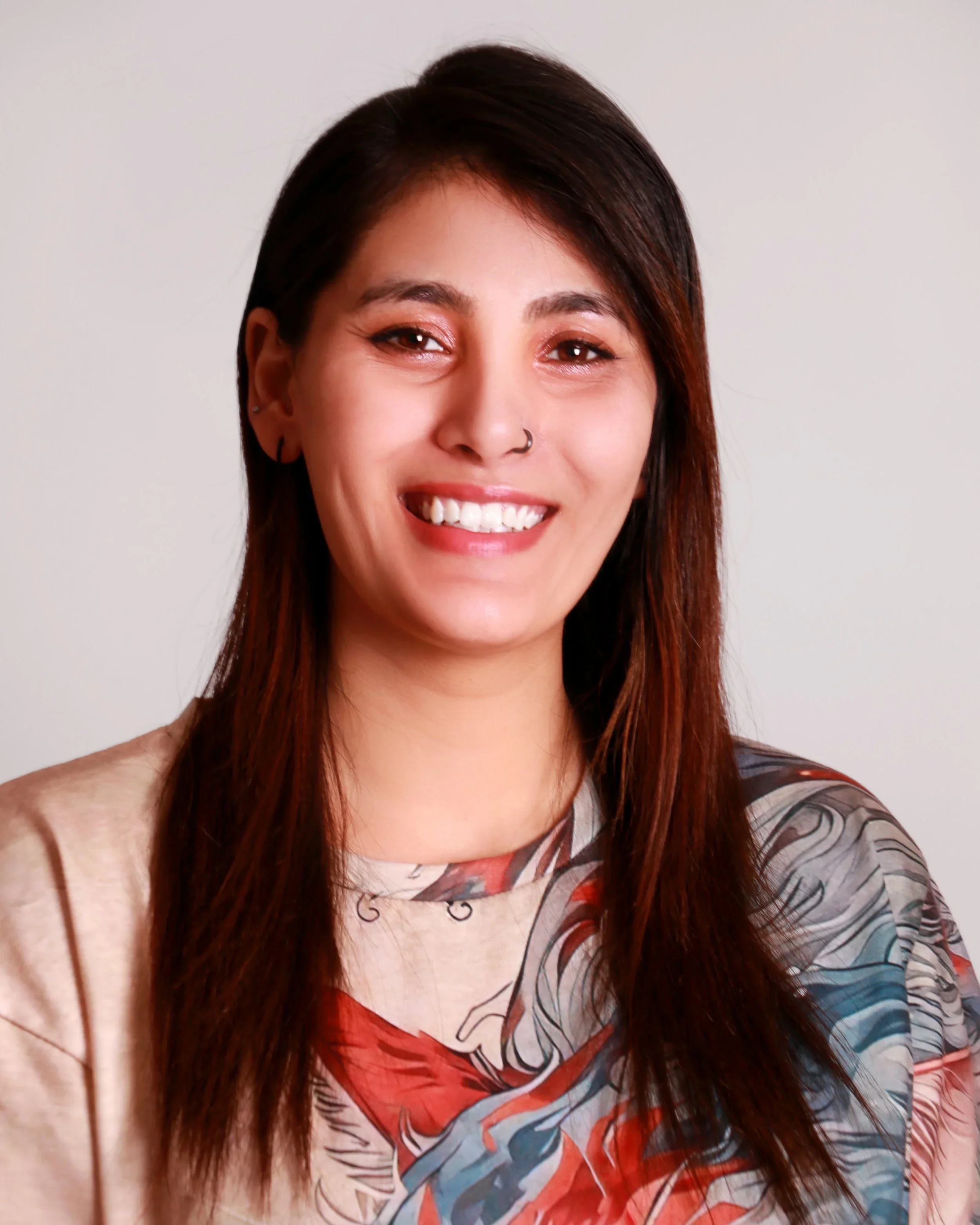 Close-up of a smiling woman with long reddish-brown hair, tattoos on her arm, wearing a colorful patterned shirt, against a plain white background.
